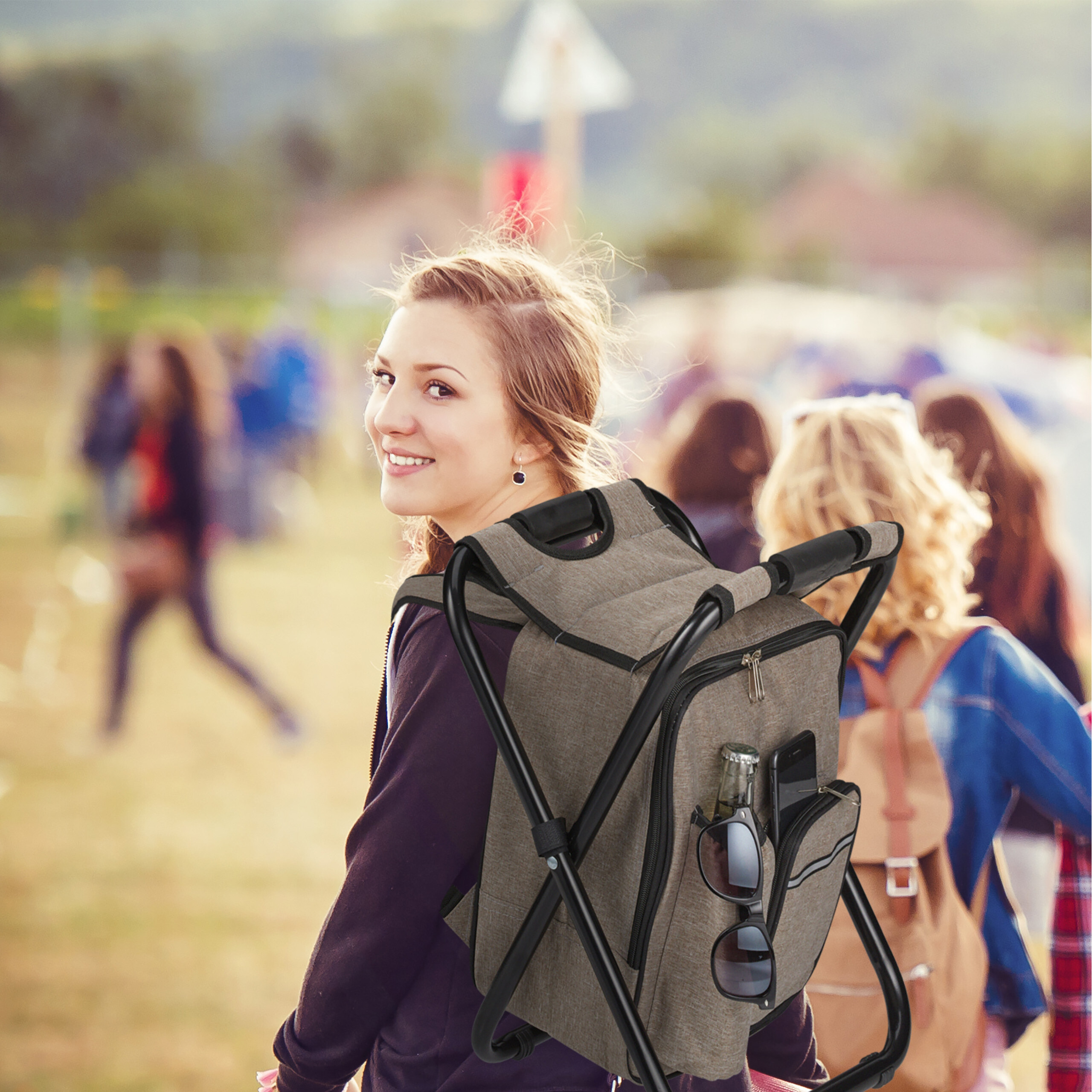 Eine junge Frau mit einem grauen Rucksack und integriertem Stuhl blickt im Freien lächelnd zurück.