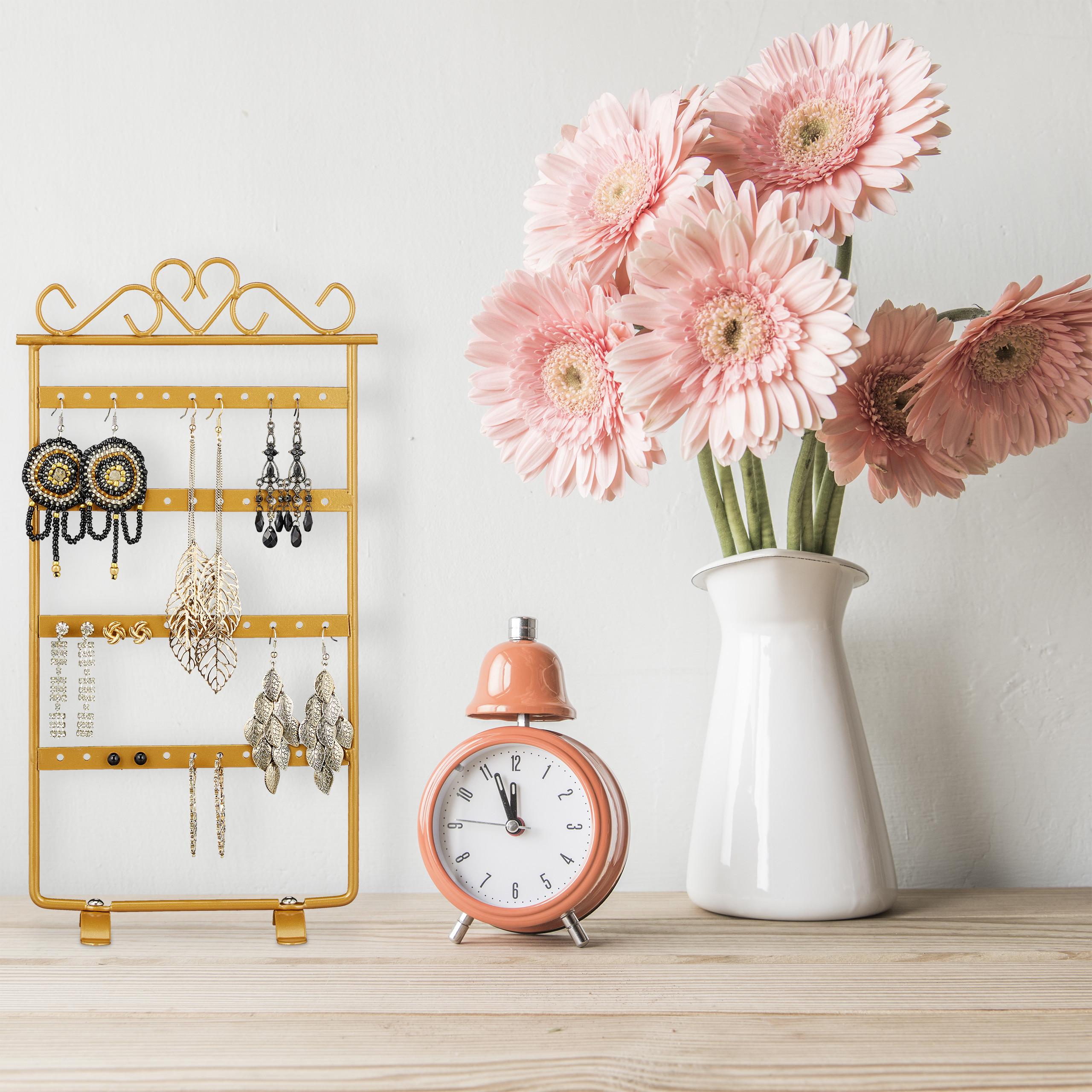 Goldener Schmuckständer, pinker Wecker und rosa Gerbera in einer weißen Vase auf Holz.