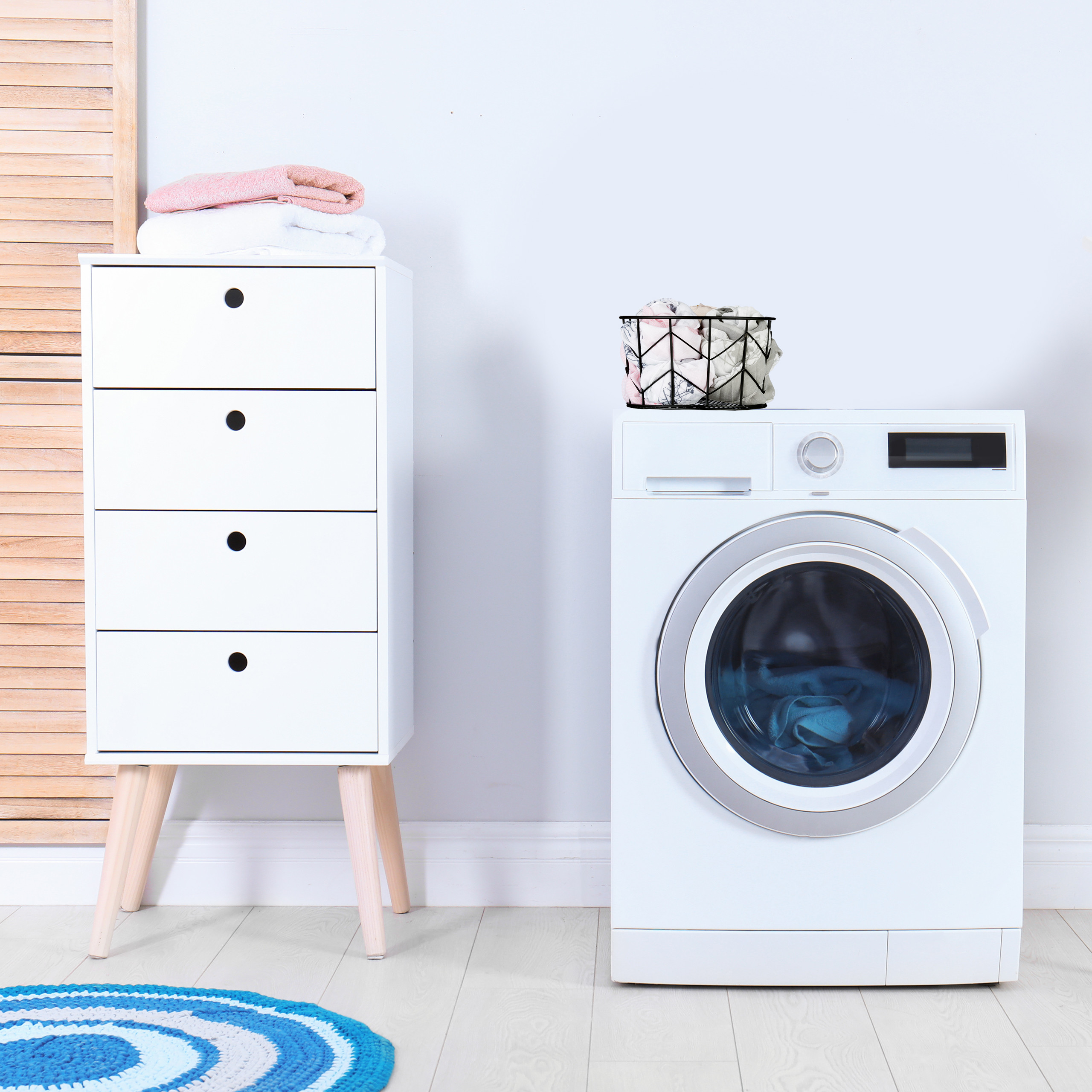 Eine weiße Waschmaschine mit blauem Teppich und weißer Vier-Schubladen-Kommode mit Handtüchern an heller Wand.