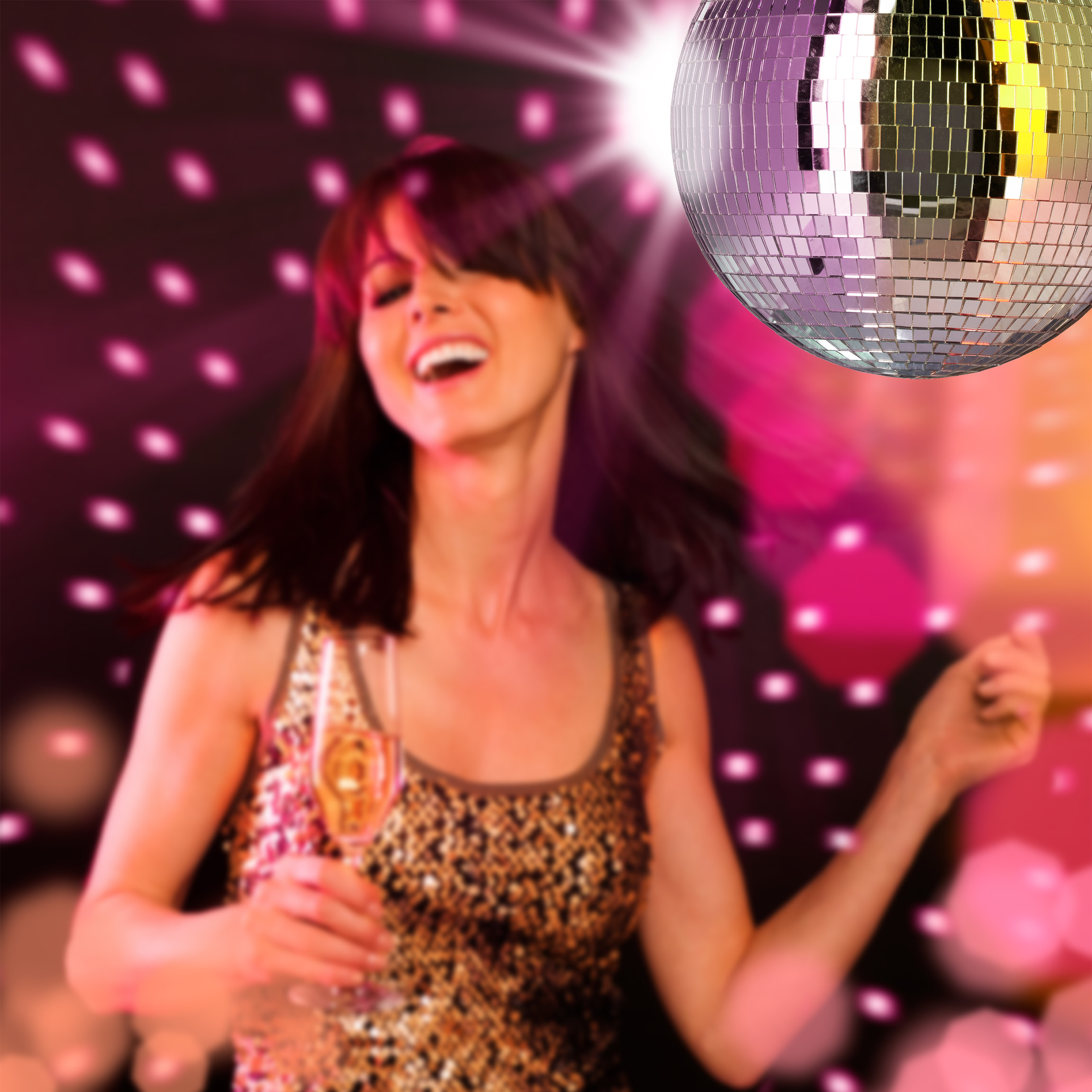 Eine Frau in einem Paillettentop hält ein Glas Sekt und tanzt unter einer Discokugel.