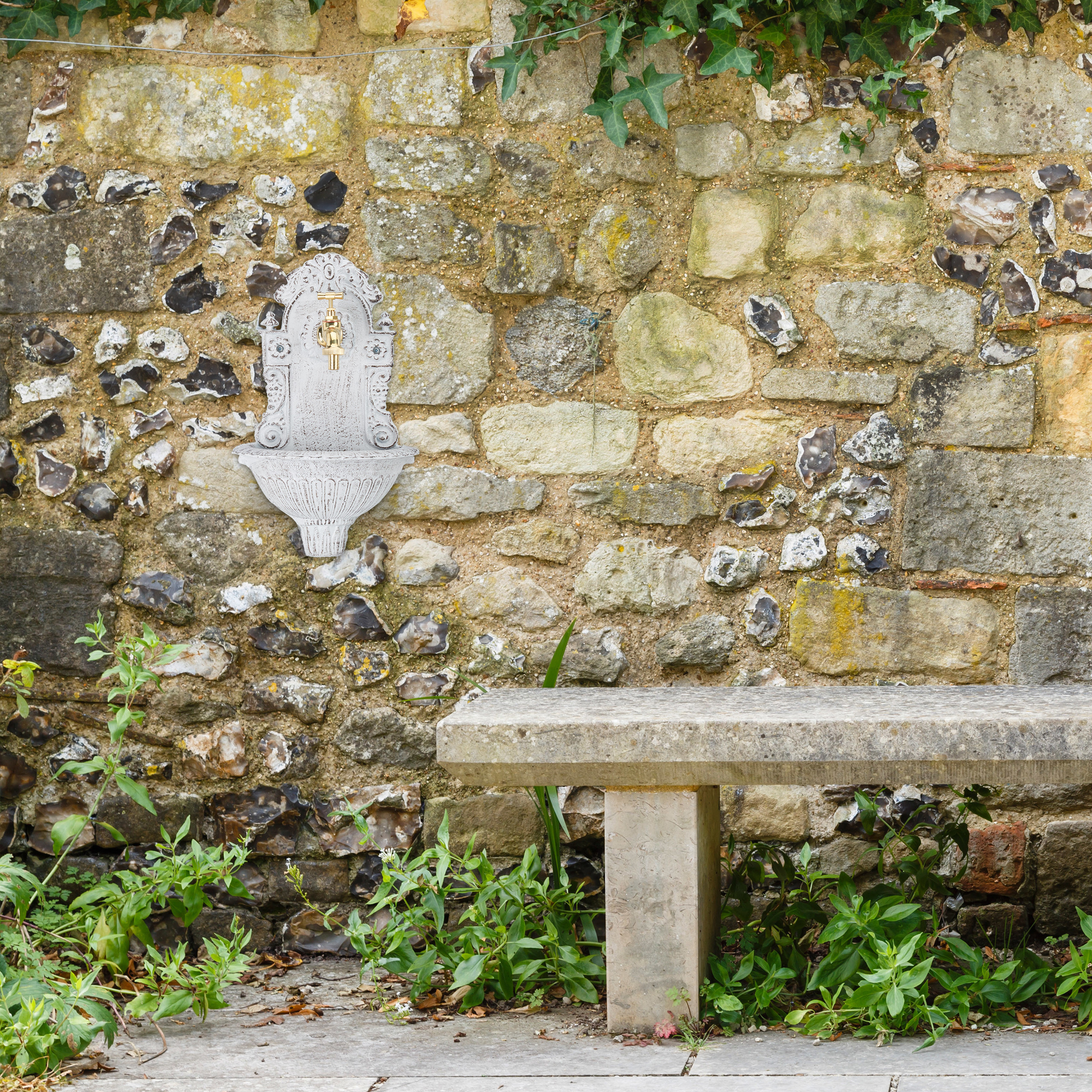 Eine Steinbank und ein weißer Wandbrunnen mit goldenem Hahn vor einer rustikalen Steinmauer mit grünen Pflanzen.