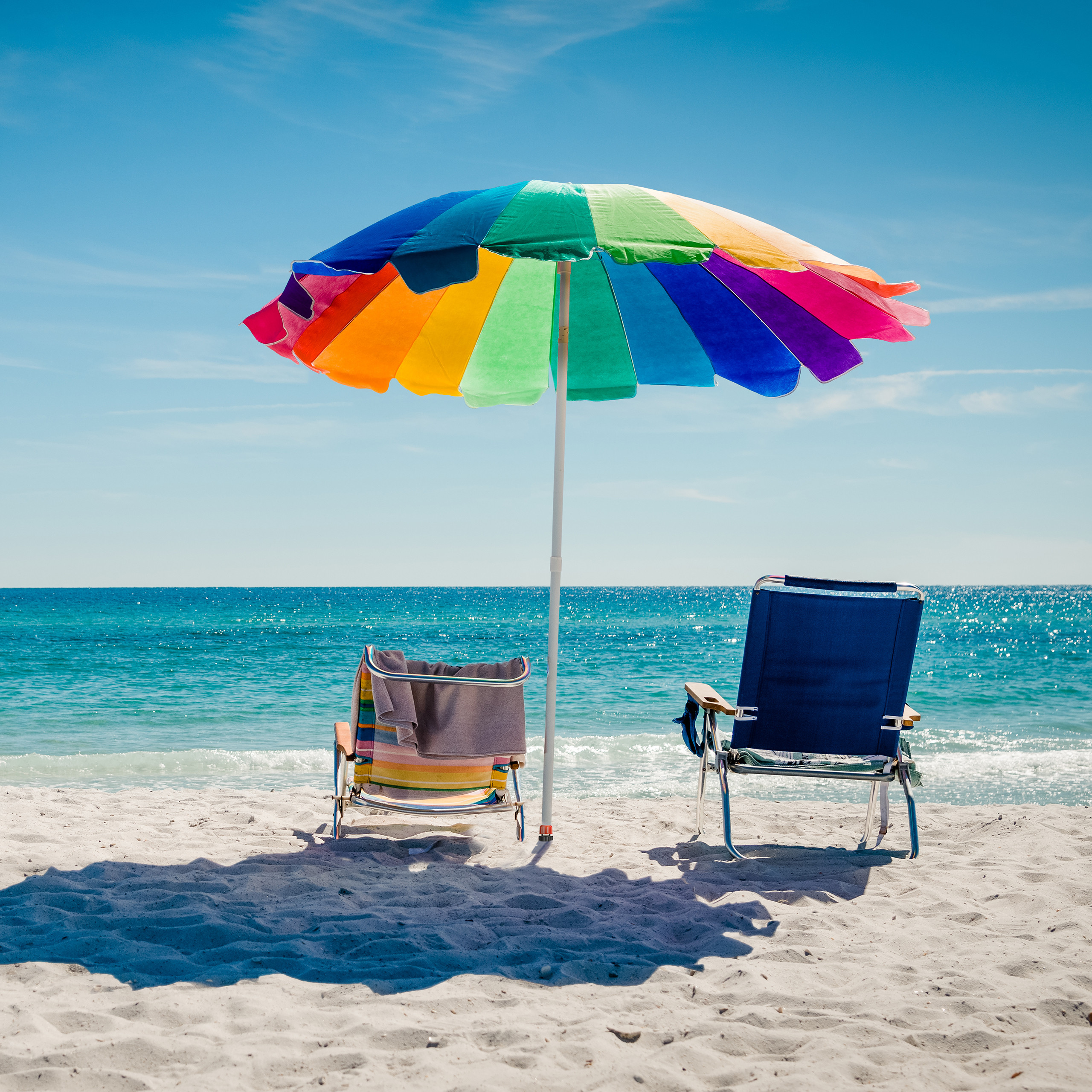 Ein bunter Sonnenschirm und zwei Strandstühle am Sandstrand mit Blick auf das blaue Meer.