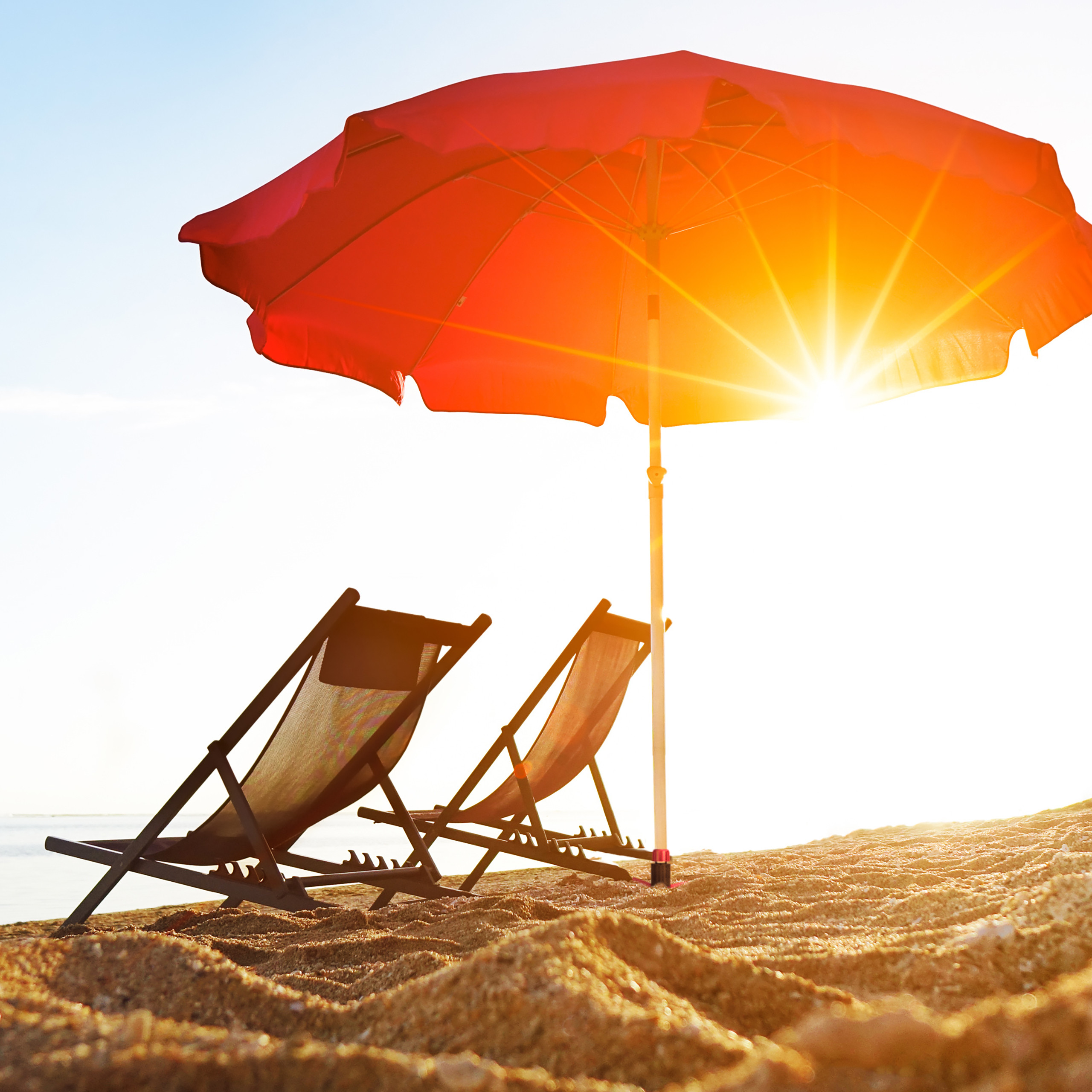 Zwei Liegestühle und ein roter Sonnenschirm am sandigen Strand mit strahlender Sonne.