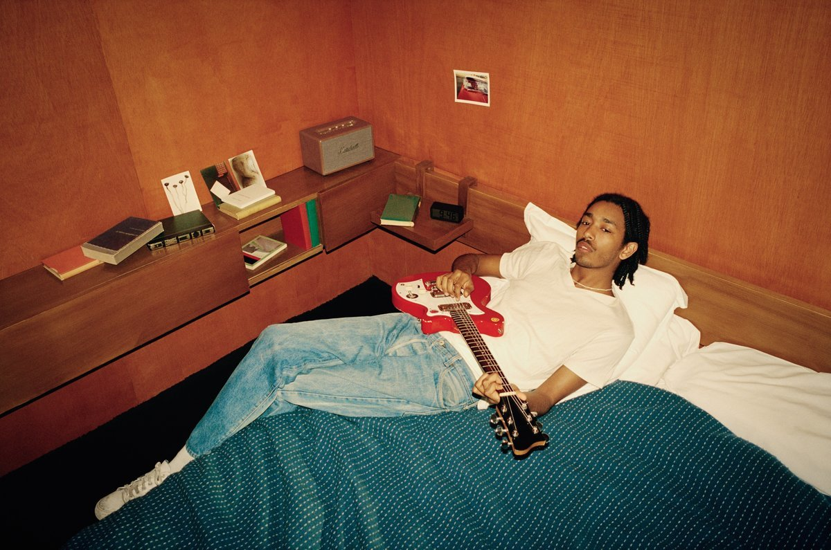 Un giovane con una chitarra è sdraiato su un letto con una coperta blu in una stanza rivestita in legno.