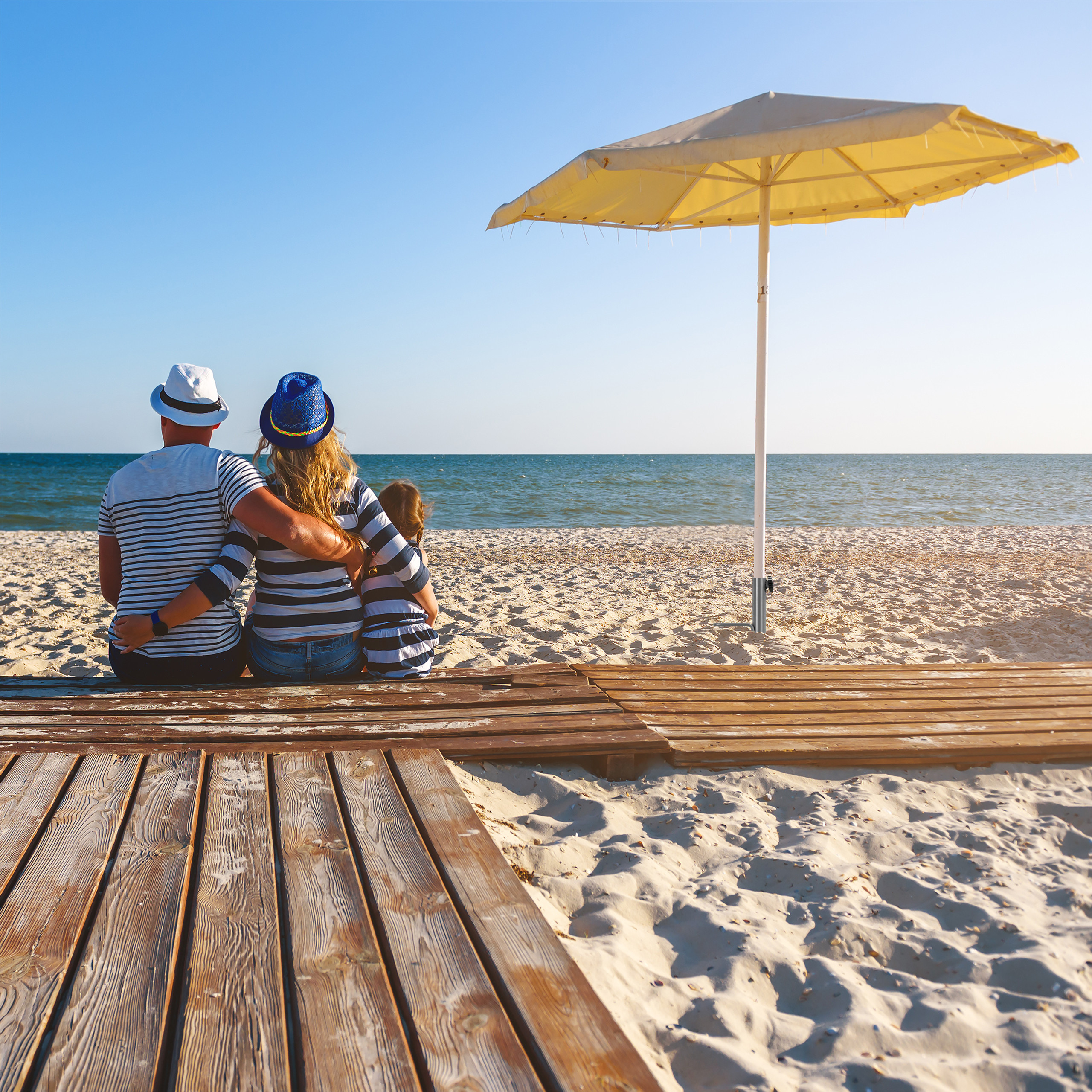 Eine Familie sitzt von hinten auf einer Holzplanke am Strand, neben einem Sonnenschirm, mit Blick auf das Meer.