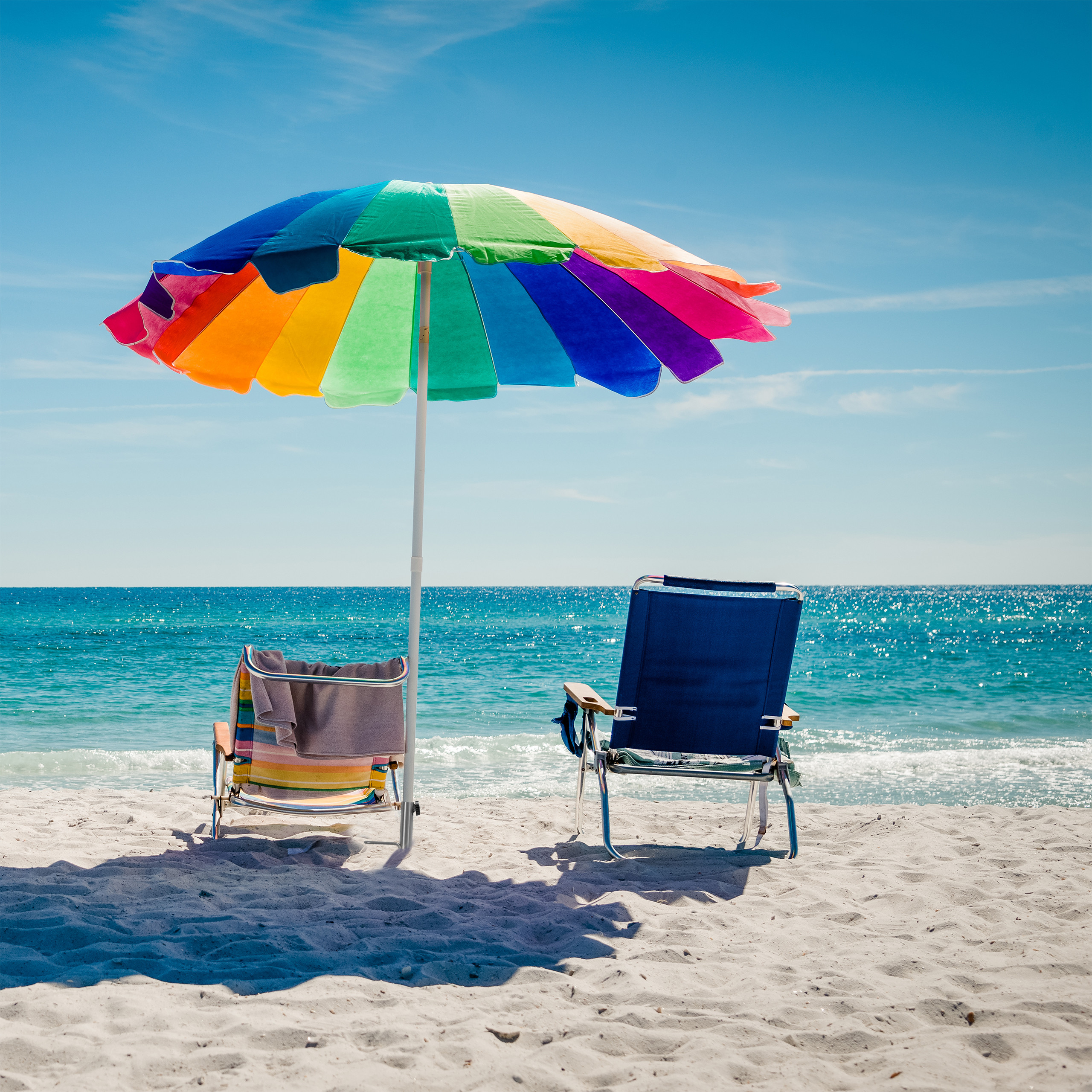 Bunter Sonnenschirm und zwei Strandstühle im weißen Sand vor dem blauen Ozean.