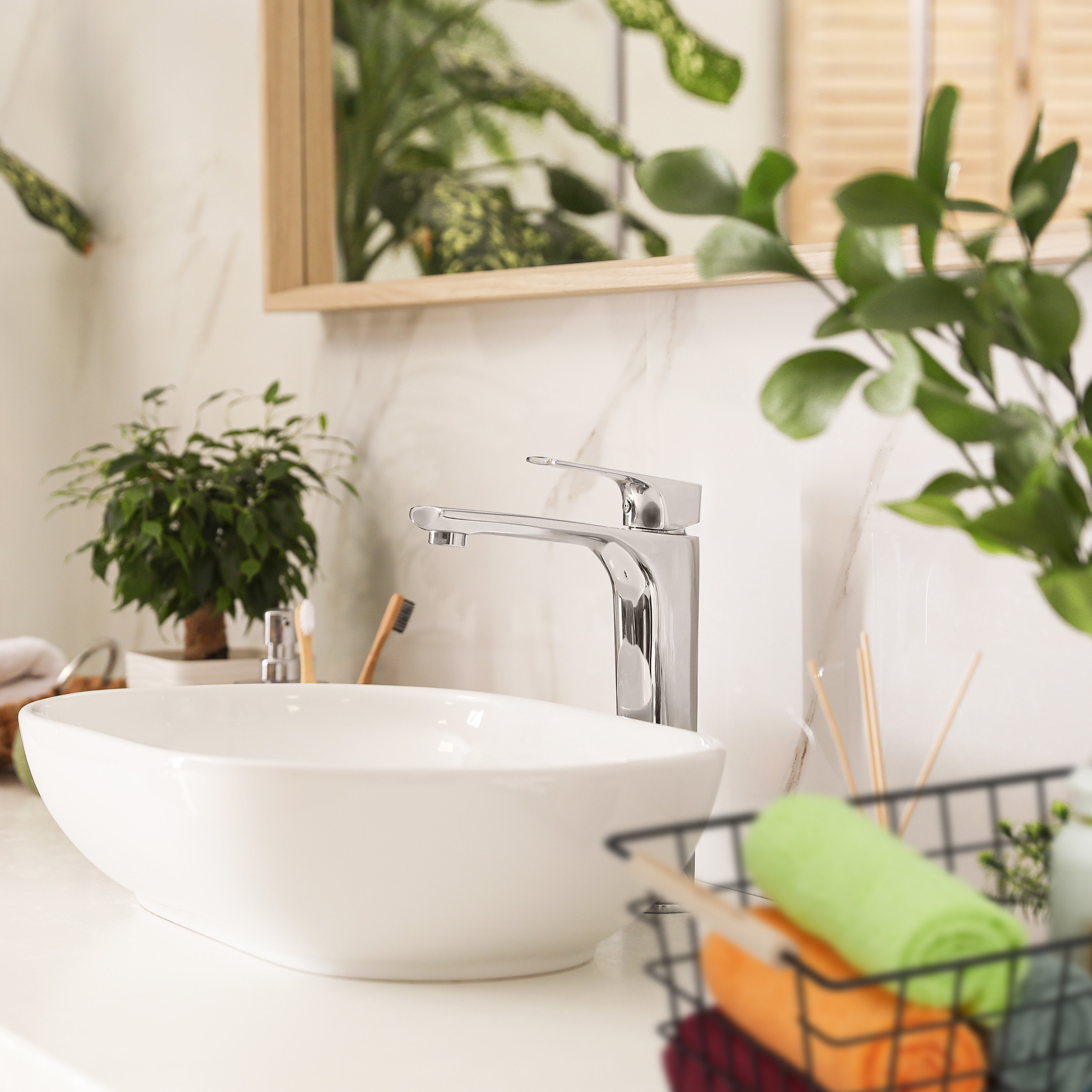 Modernes weißes Waschbecken mit Chromarmatur auf einer Arbeitsplatte, umgeben von grünen Pflanzen und Badezimmerzubehör.