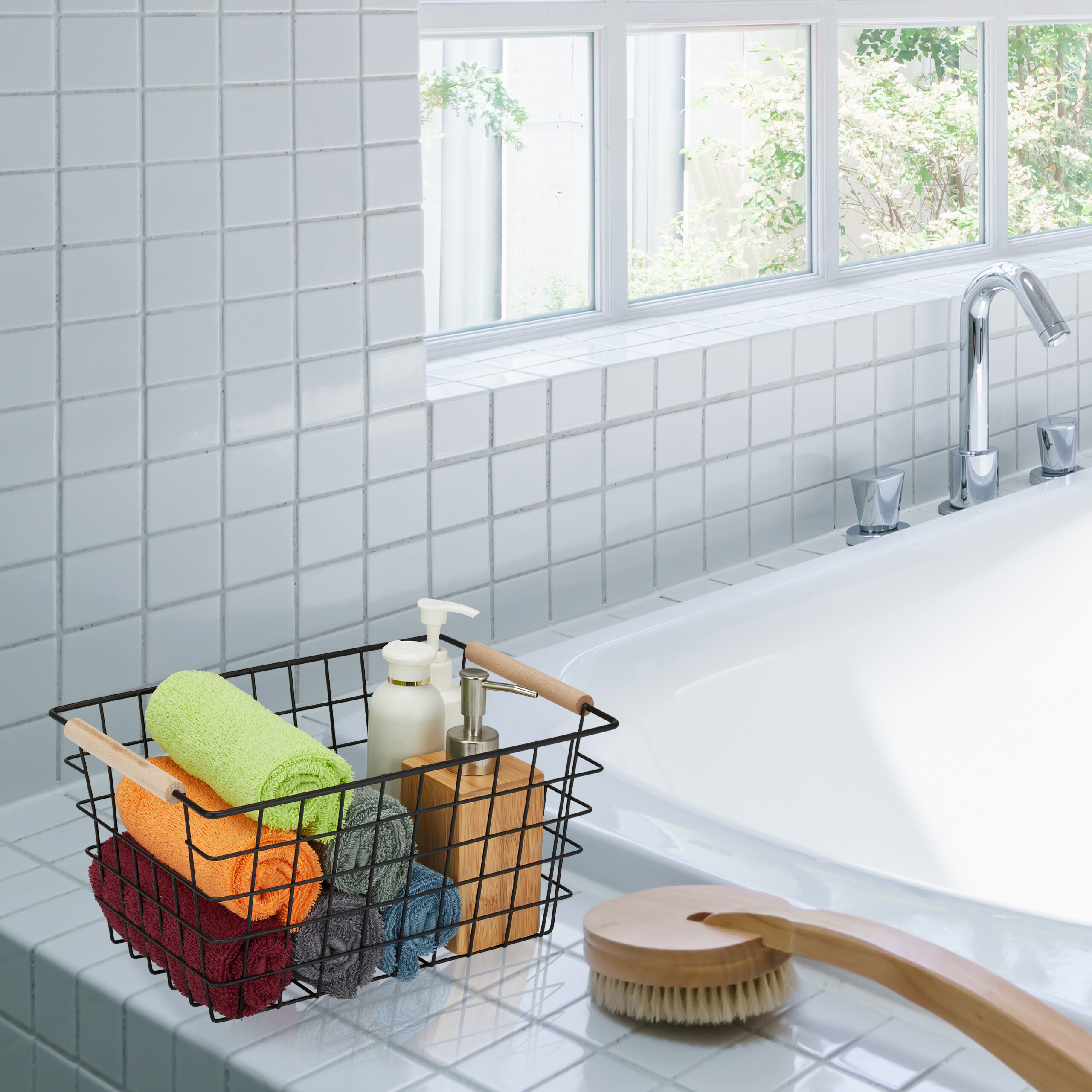 Ein Drahtkorb mit bunten Handtüchern und Seifenspendern steht auf dem gefliesten Rand einer Badewanne.