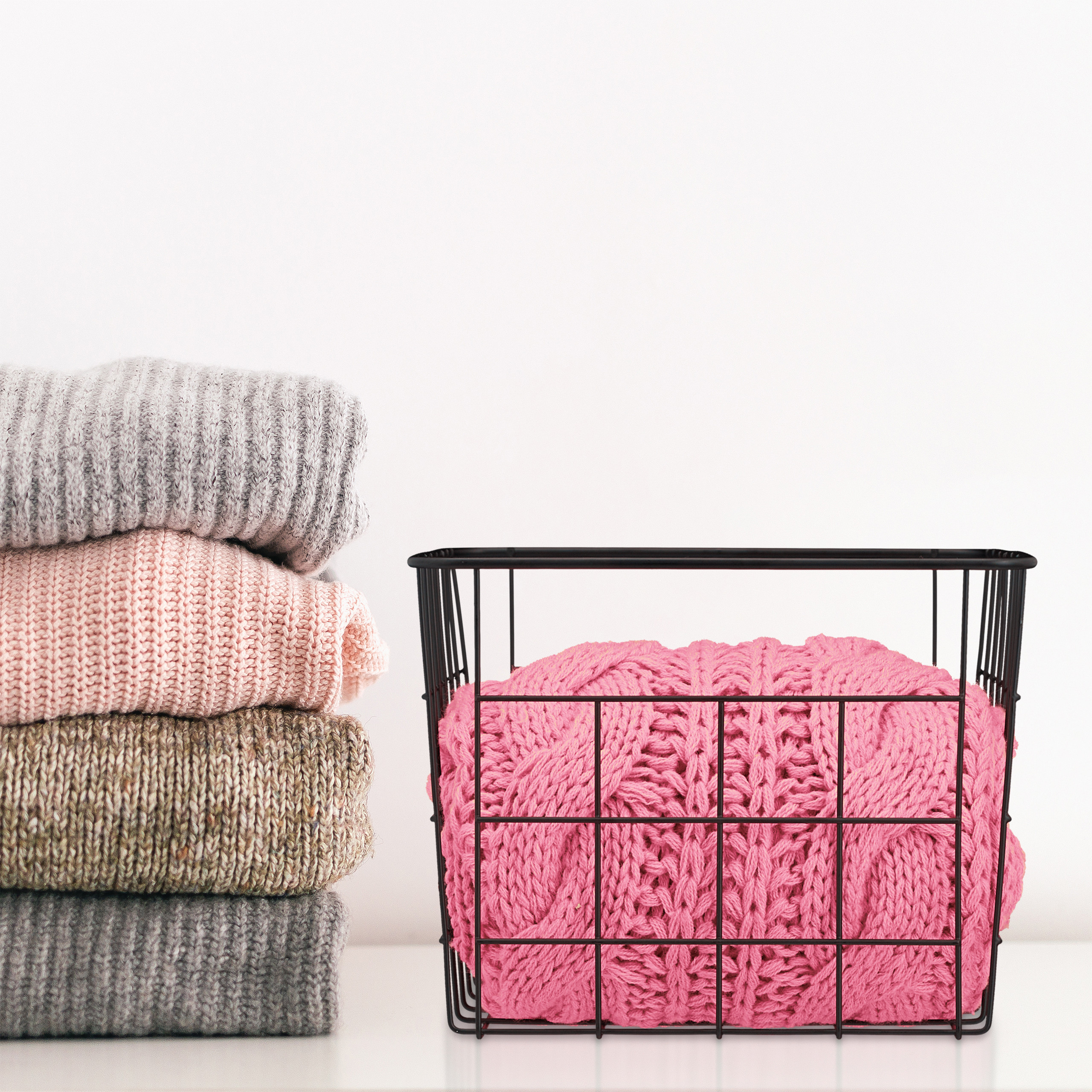 Ein Stapel Strickpullover in Grau, Rosa und Braun steht neben einem schwarzen Drahtkorb mit einer rosa Decke.