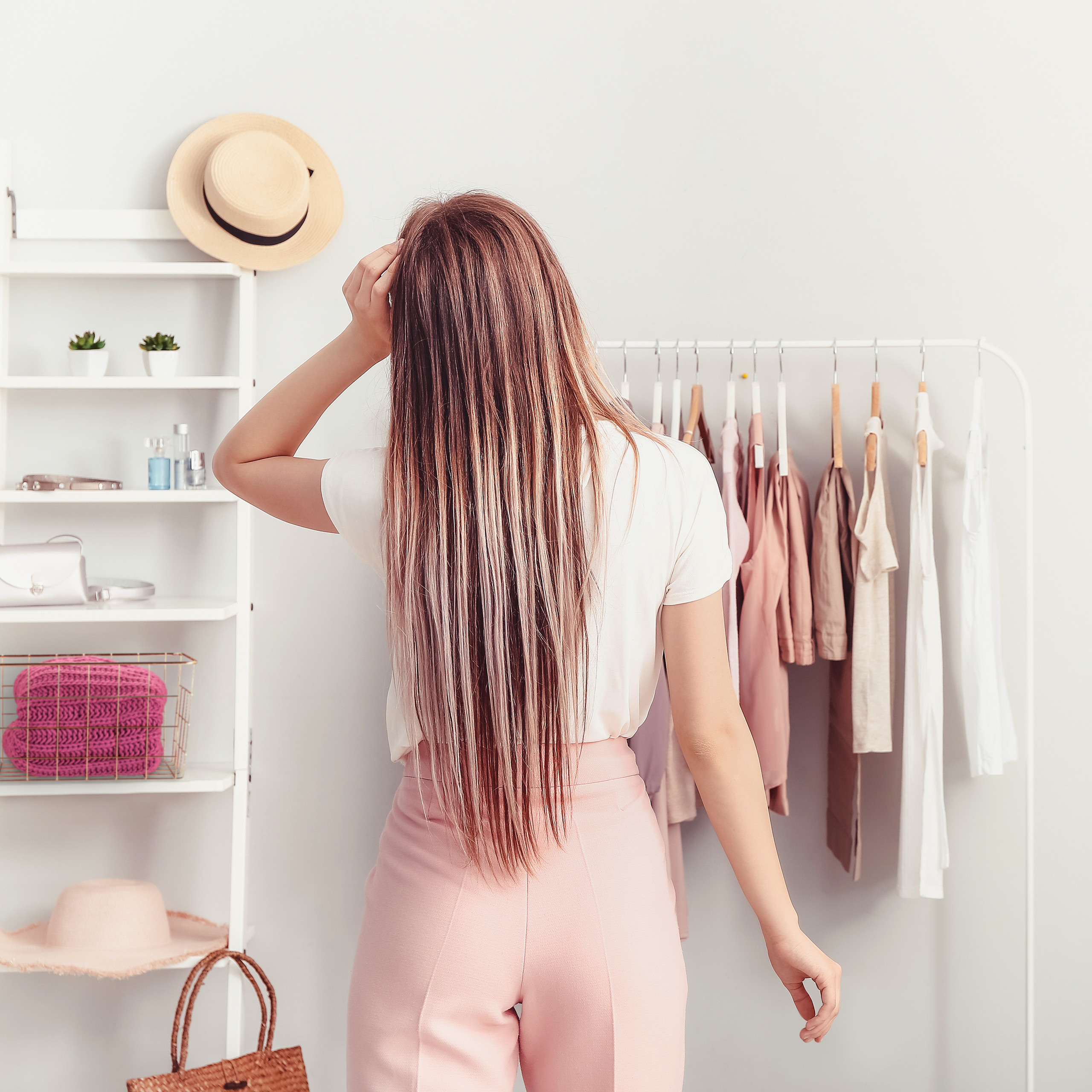 Eine langhaarige Frau in weißem Top und pinken Hosen steht vor einem Kleiderständer und Regalen in einem Raum.