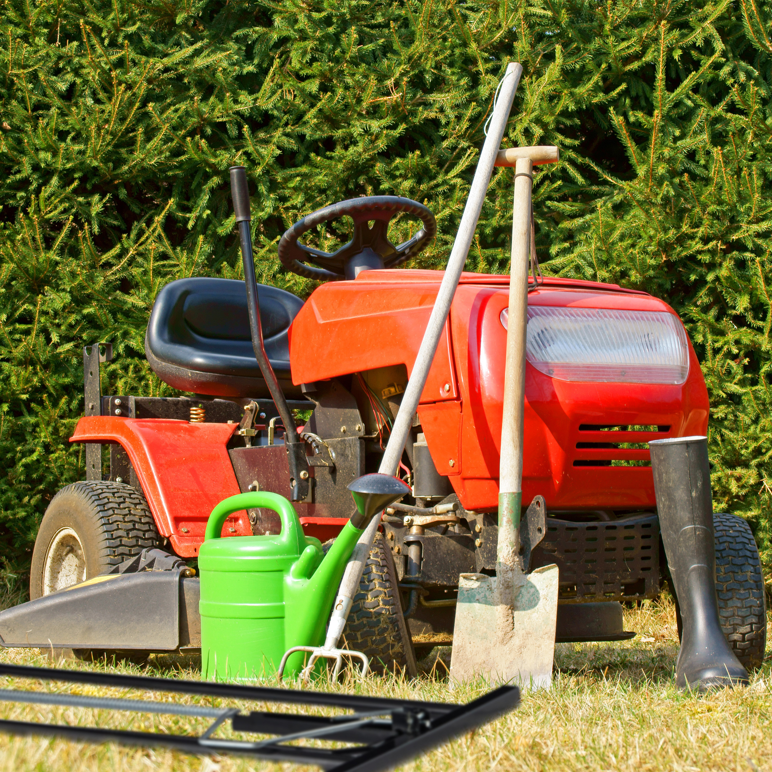 Roter Aufsitzrasenmäher mit grüner Gießkanne, Schaufel und Stiefel vor einer Hecke.