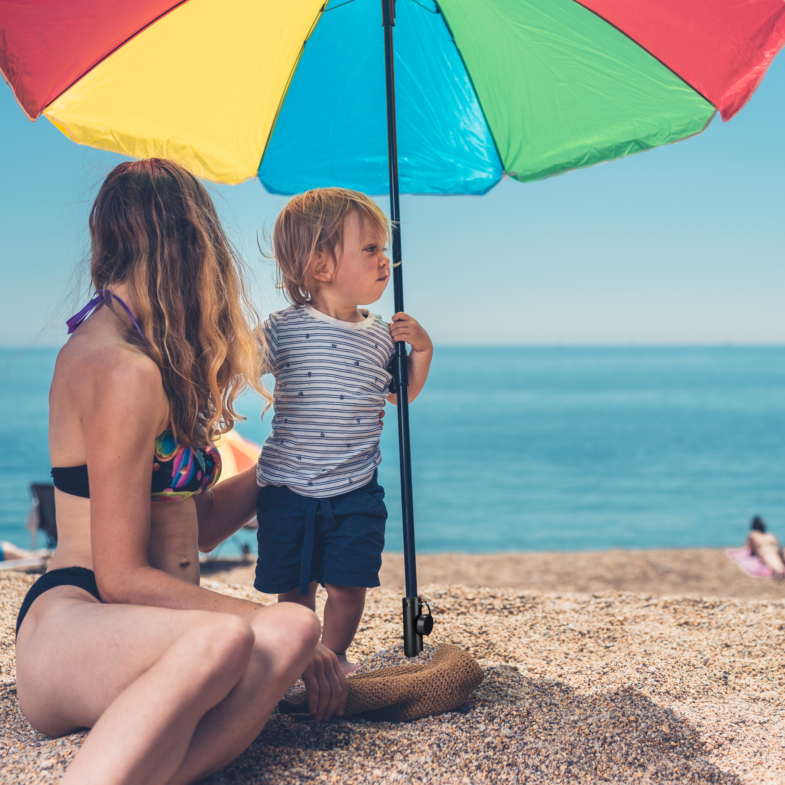 Eine Frau in Bikini und ein Kind sitzen am Strand unter einem bunten Sonnenschirm.