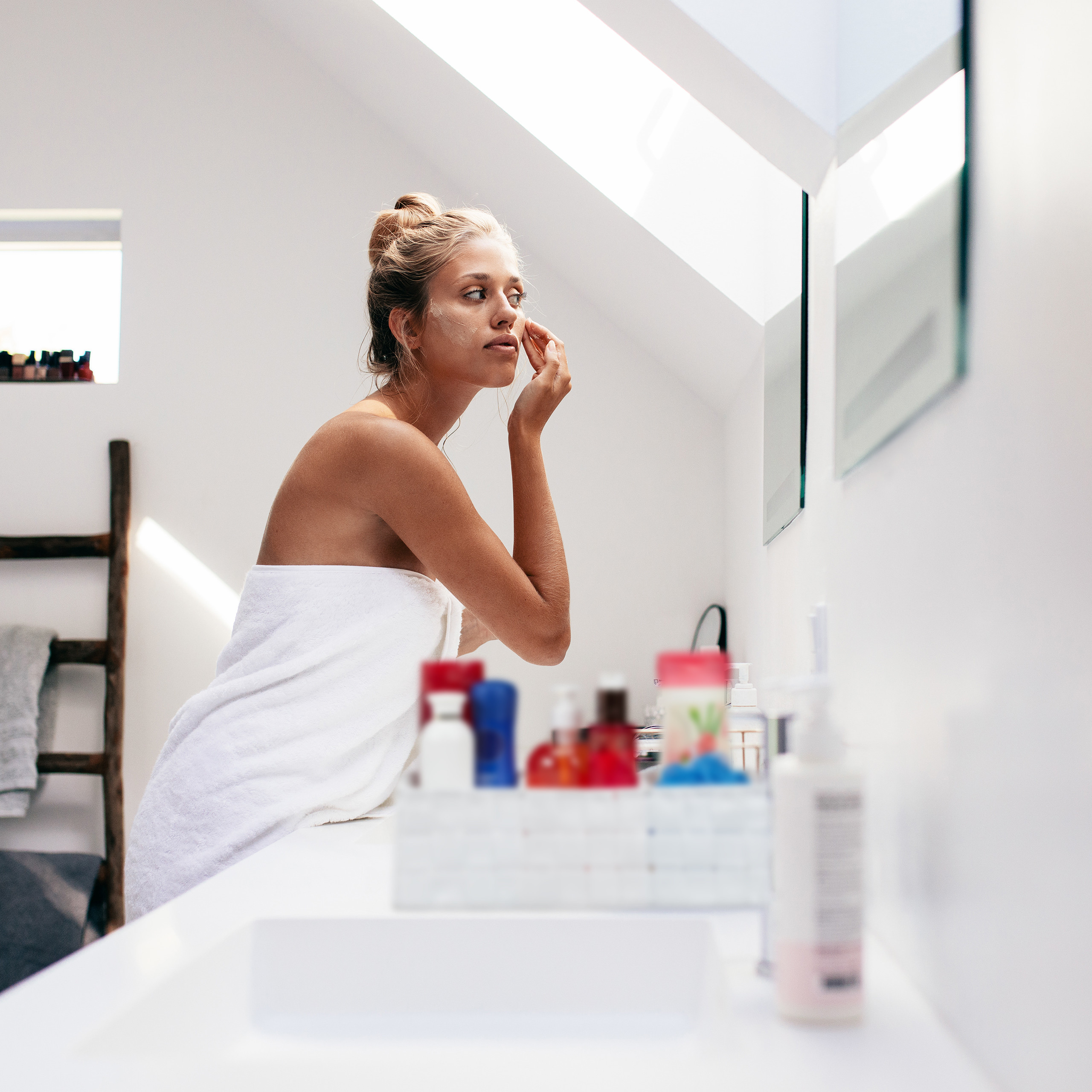Eine blonde Frau in einem Handtuch steht vor einem Badezimmerspiegel und trägt Gesichtscreme auf.