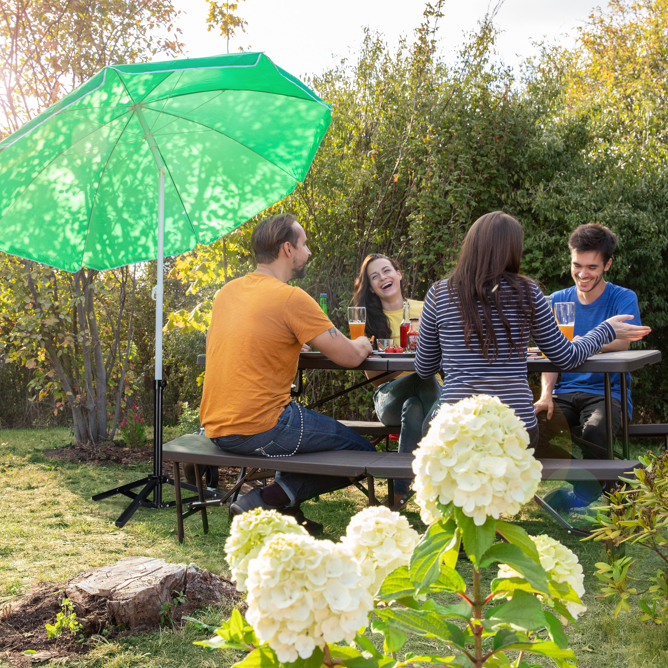 Vier Freunde sitzen unter einem großen grünen Sonnenschirm an einem Picknicktisch im Garten.