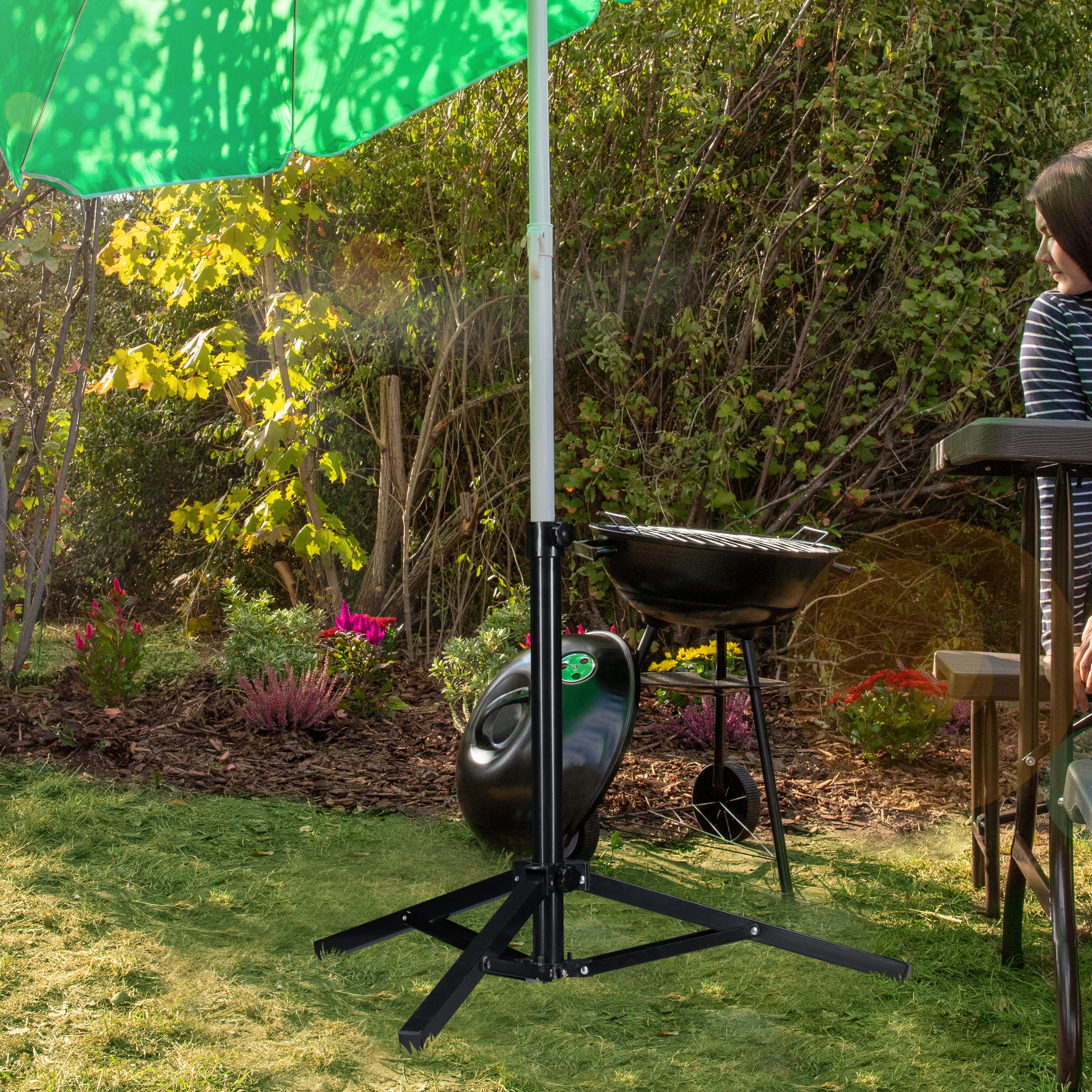 Grüner Sonnenschirm mit Ständer im Garten, daneben ein Holzkohlegrill und eine Frau an einem Tisch.