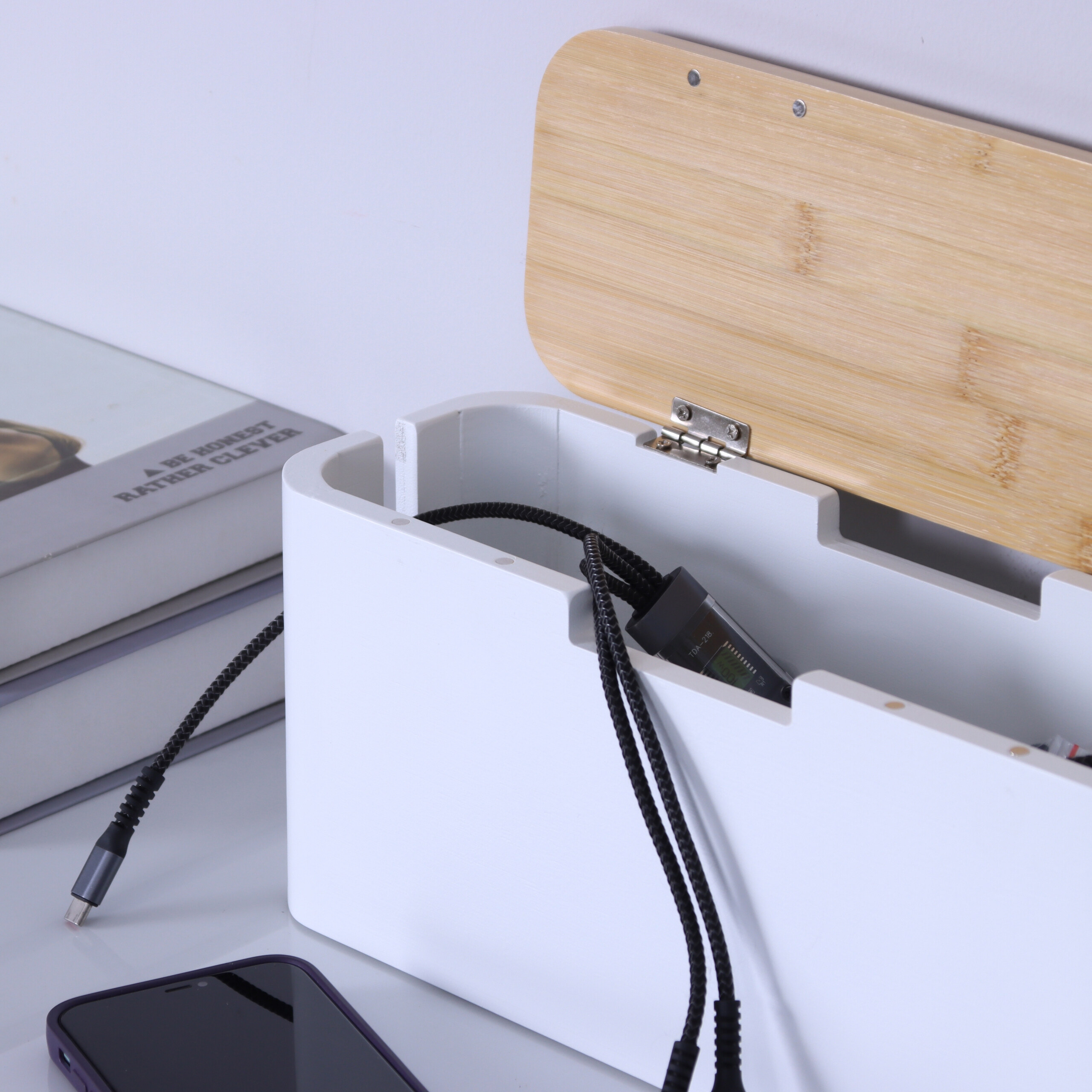 Eine weiße Kabelbox mit offenem Holzdeckel, die schwarze Kabel und ein Ladegerät enthält.