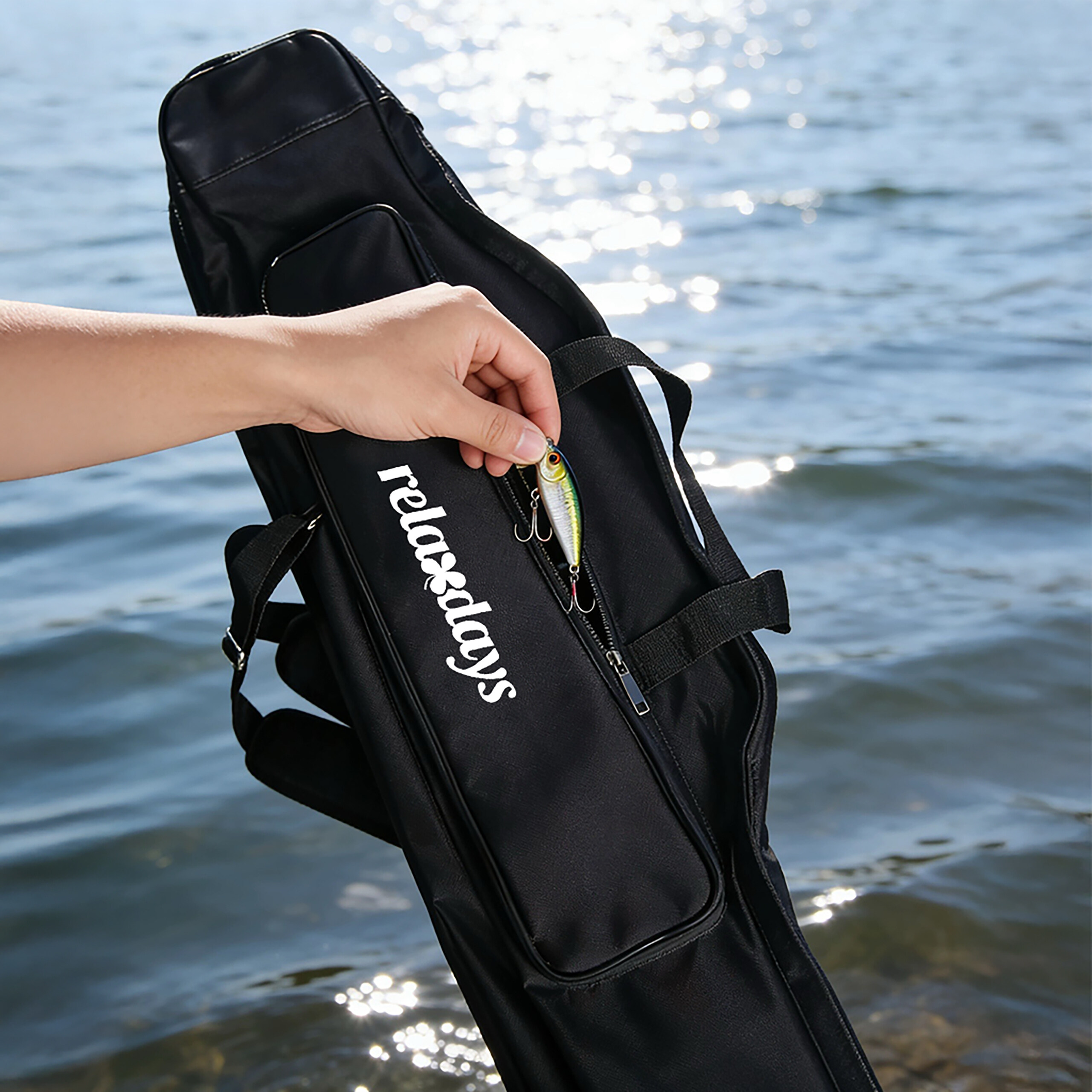Eine Hand greift nach einem Angelköder an einer schwarzen Angeltasche vor glitzerndem Wasser.