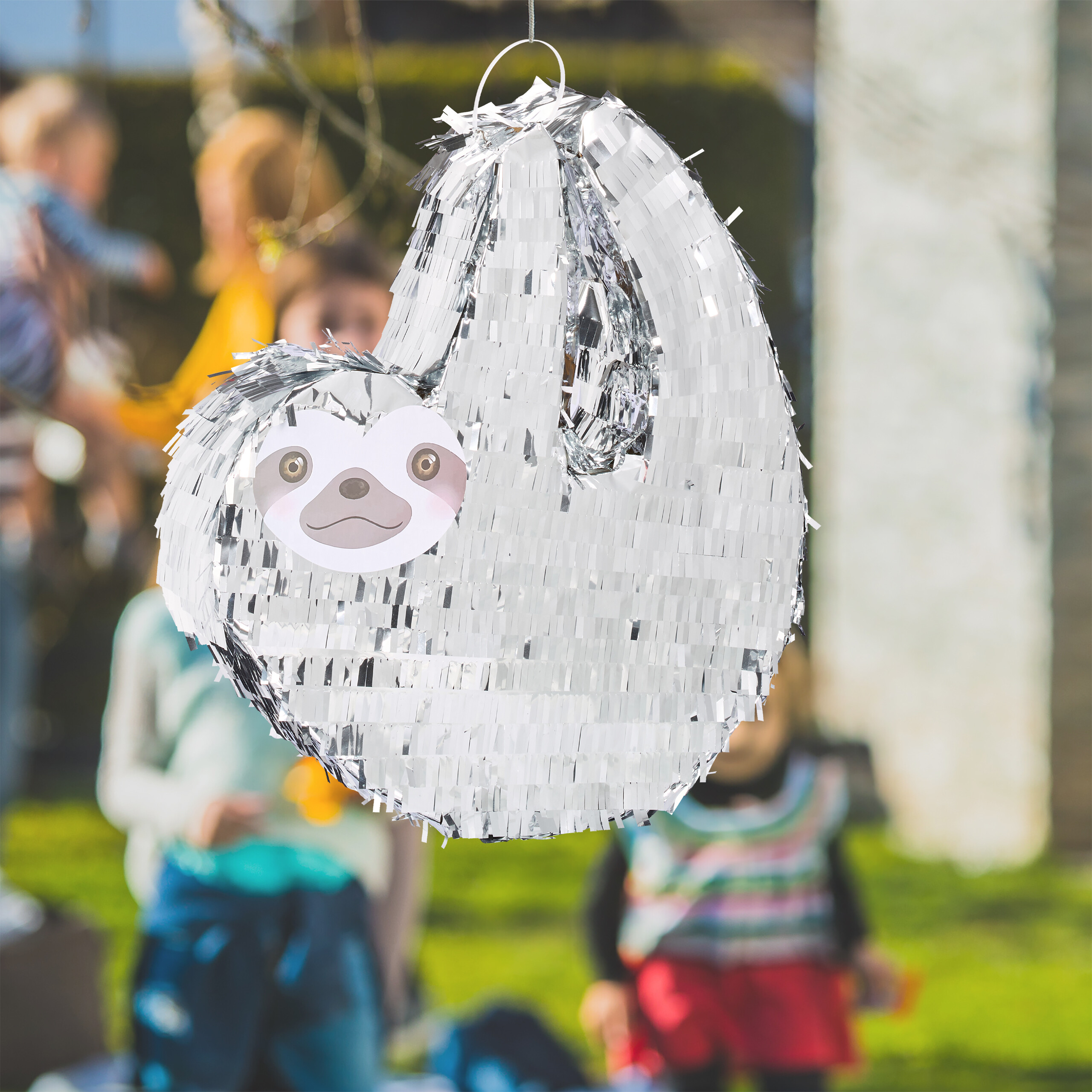 Eine silberne Faultier-Piñata hängt draußen im Garten, mit verschwommenen Personen im Hintergrund.