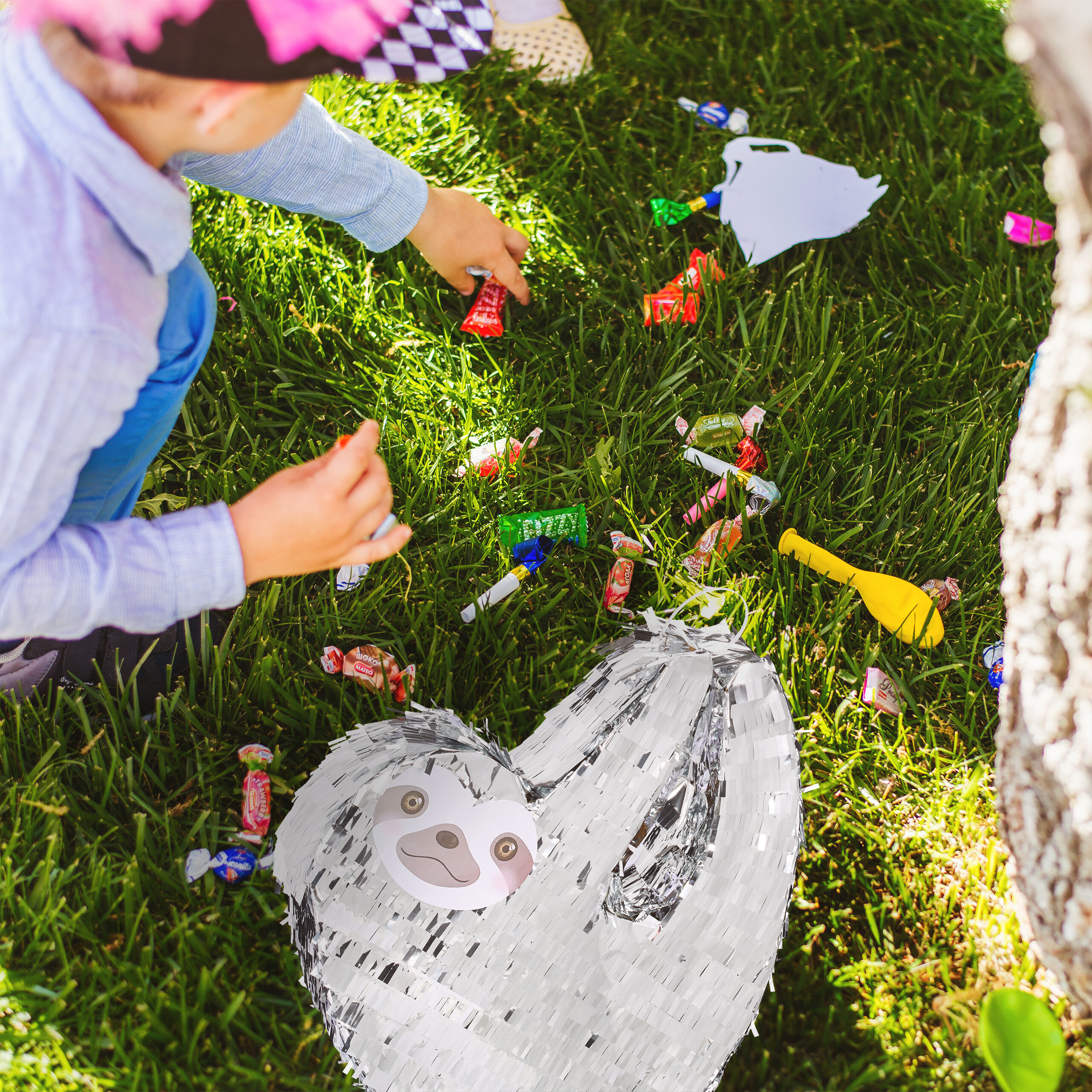 Ein Kind im blauen Hemd hebt Süßigkeiten auf dem grünen Gras neben einer silbernen Faultier-Piñata auf.