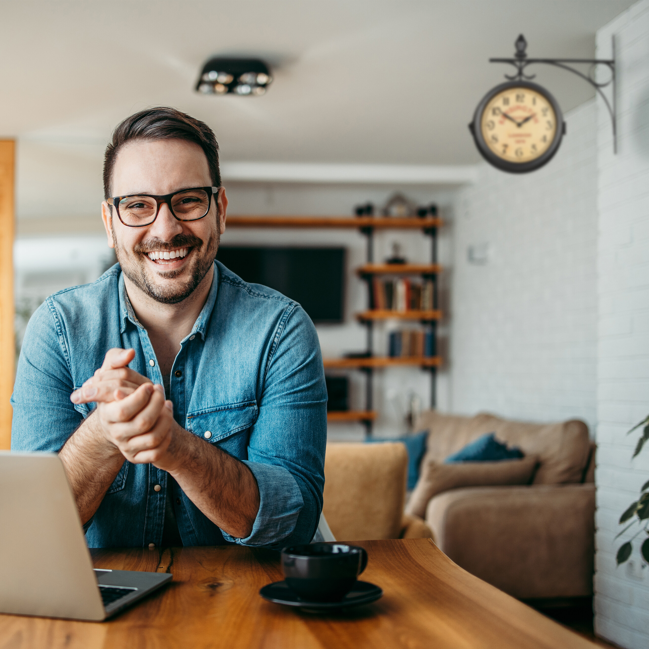 Ein lächelnder Mann mit Brille sitzt an einem Holztisch mit Laptop und Kaffeetasse in einer hellen Wohnung.