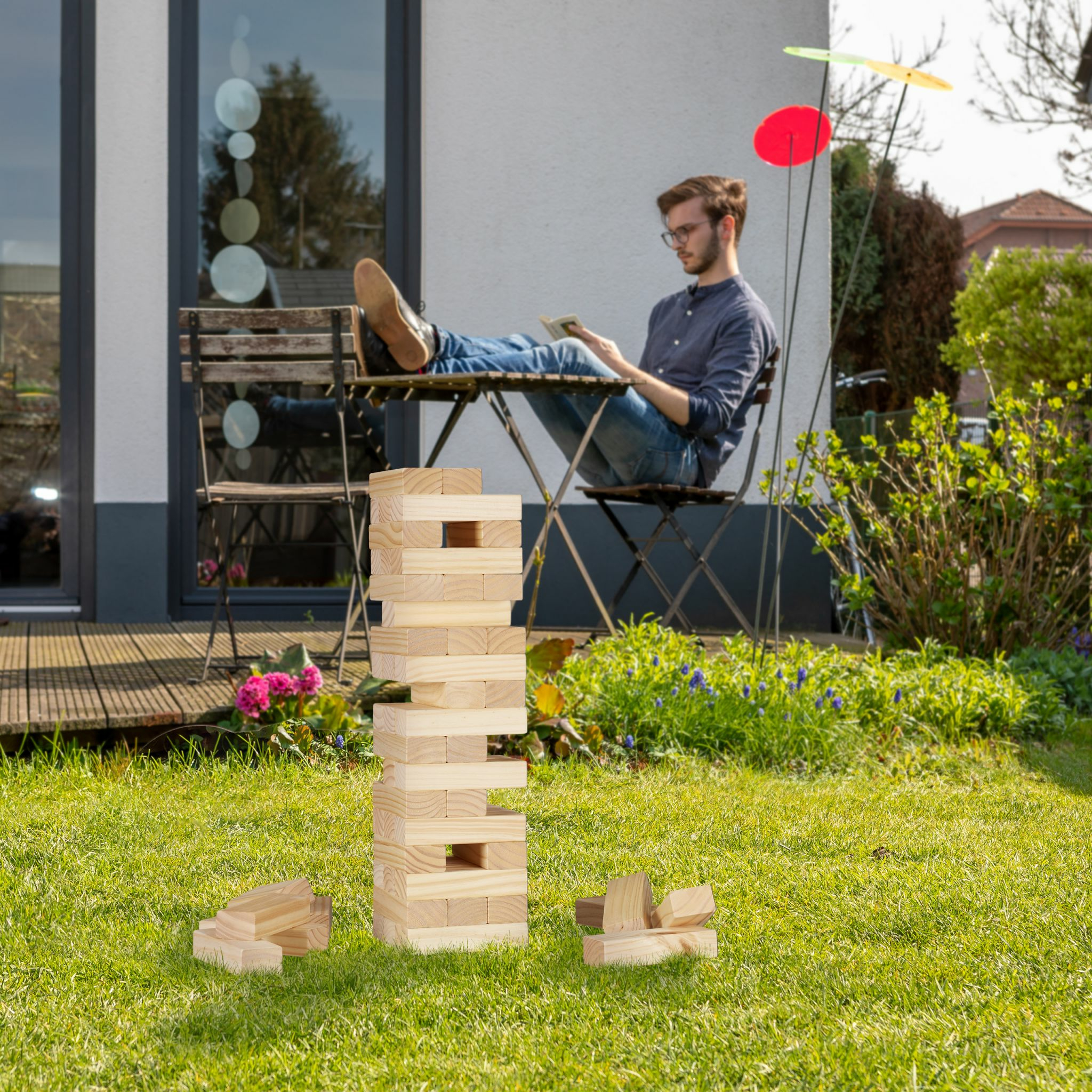Ein Jenga-Turm steht auf dem Gras, während ein Mann im Hintergrund am Tisch sitzt und liest.