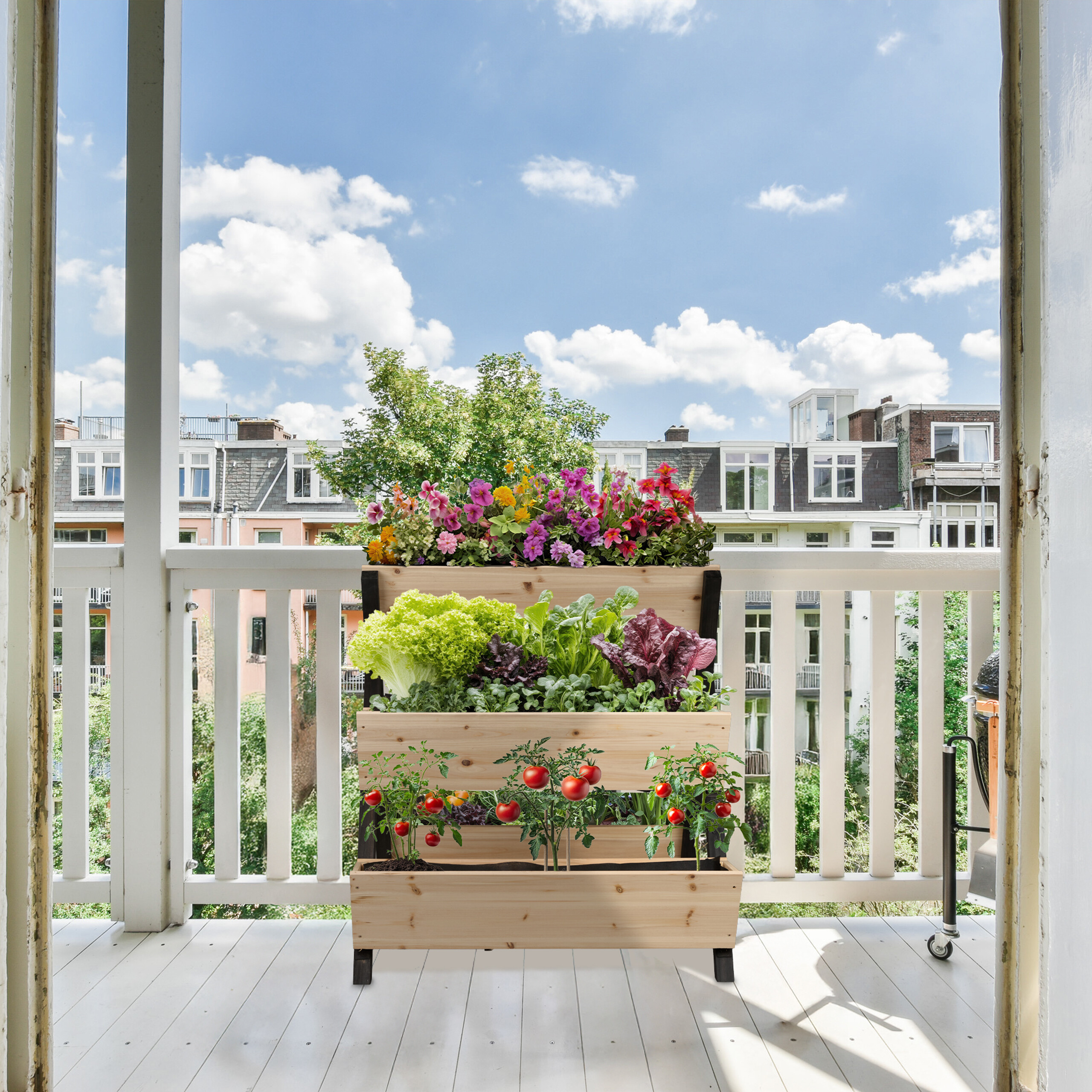 Ein hölzernes Hochbeet auf einem sonnigen Balkon mit Blumen, Salat und Tomatenpflanzen vor Stadtgebäuden.