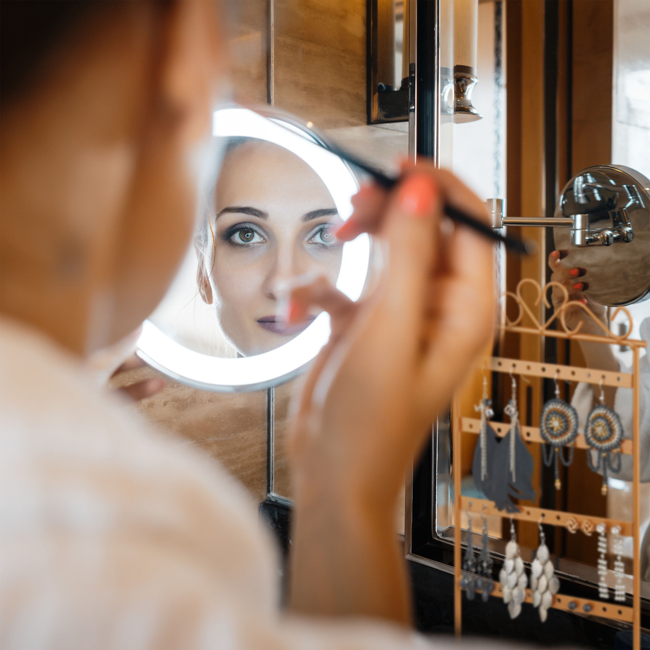 Frau mit Make-up-Pinsel blickt in einen beleuchteten Kosmetikspiegel.