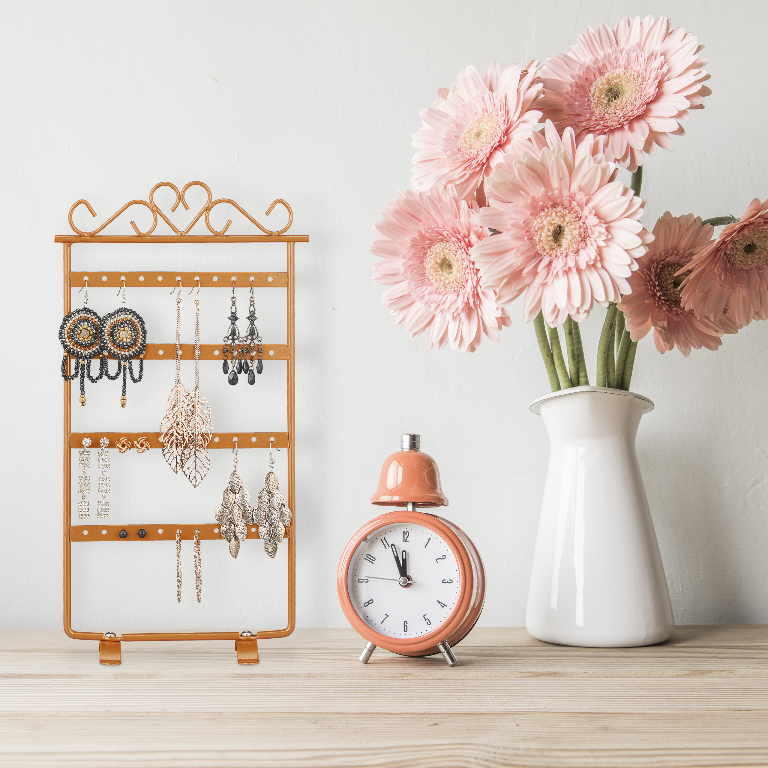 Ein goldener Schmuckständer, ein orangefarbener Wecker und rosa Gerbera in einer weißen Vase.