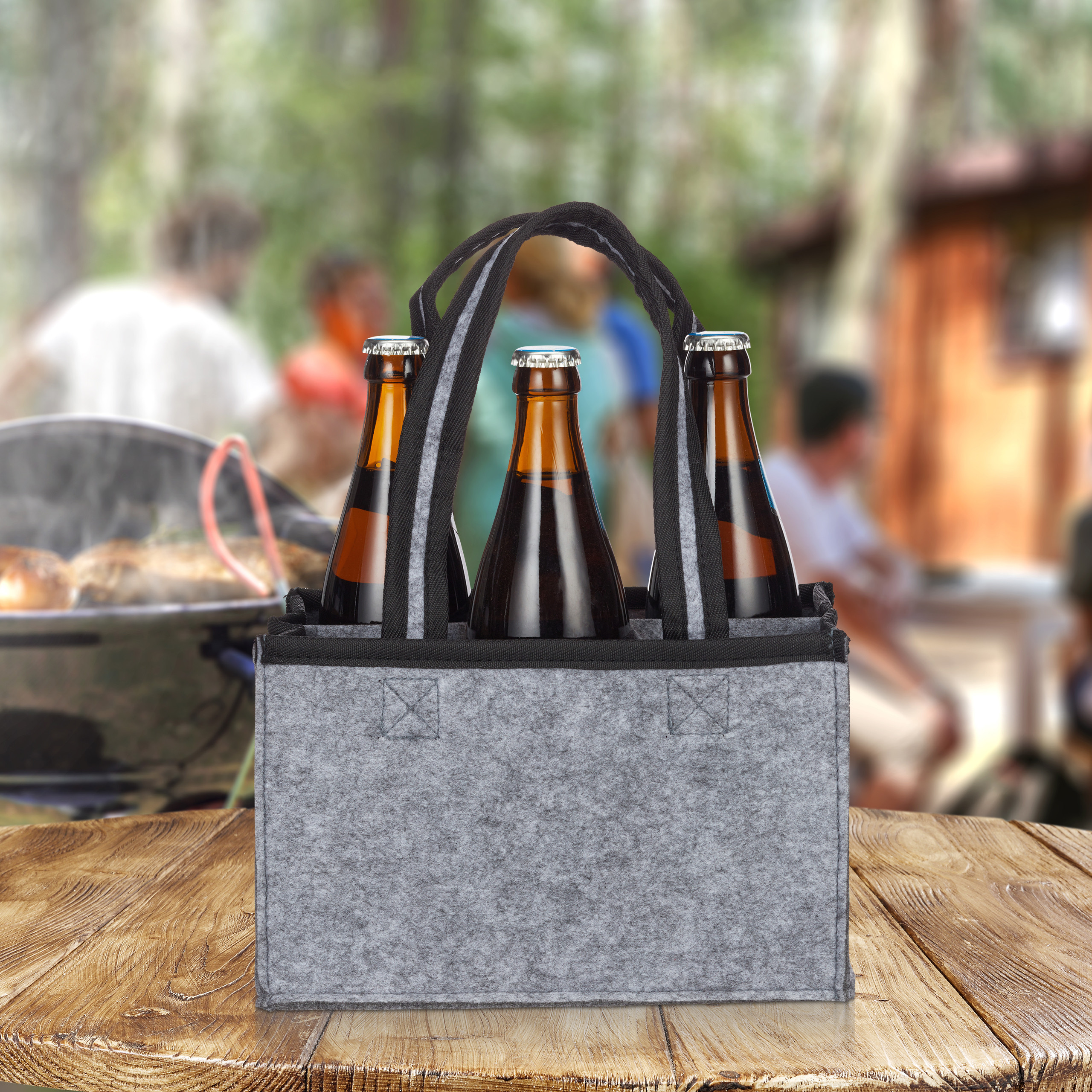 Ein grauer Filzkorb mit drei braunen Bierflaschen auf einem Holztisch vor verschwommenem Grillhintergrund.