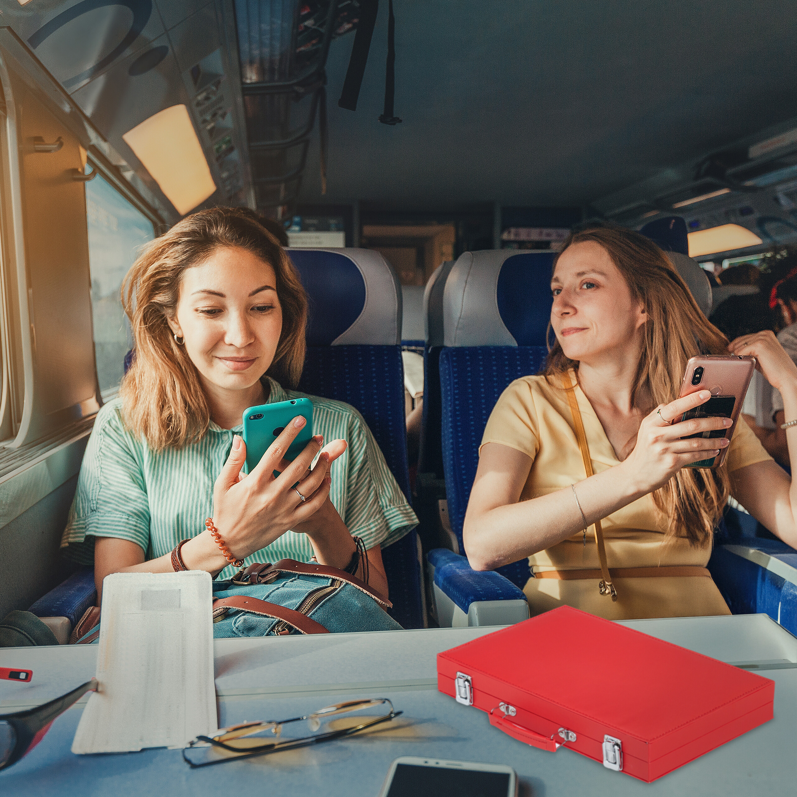Zwei Frauen sitzen im Zug, nutzen Smartphones und blicken auf ein kleines rotes Etui auf dem Tisch.