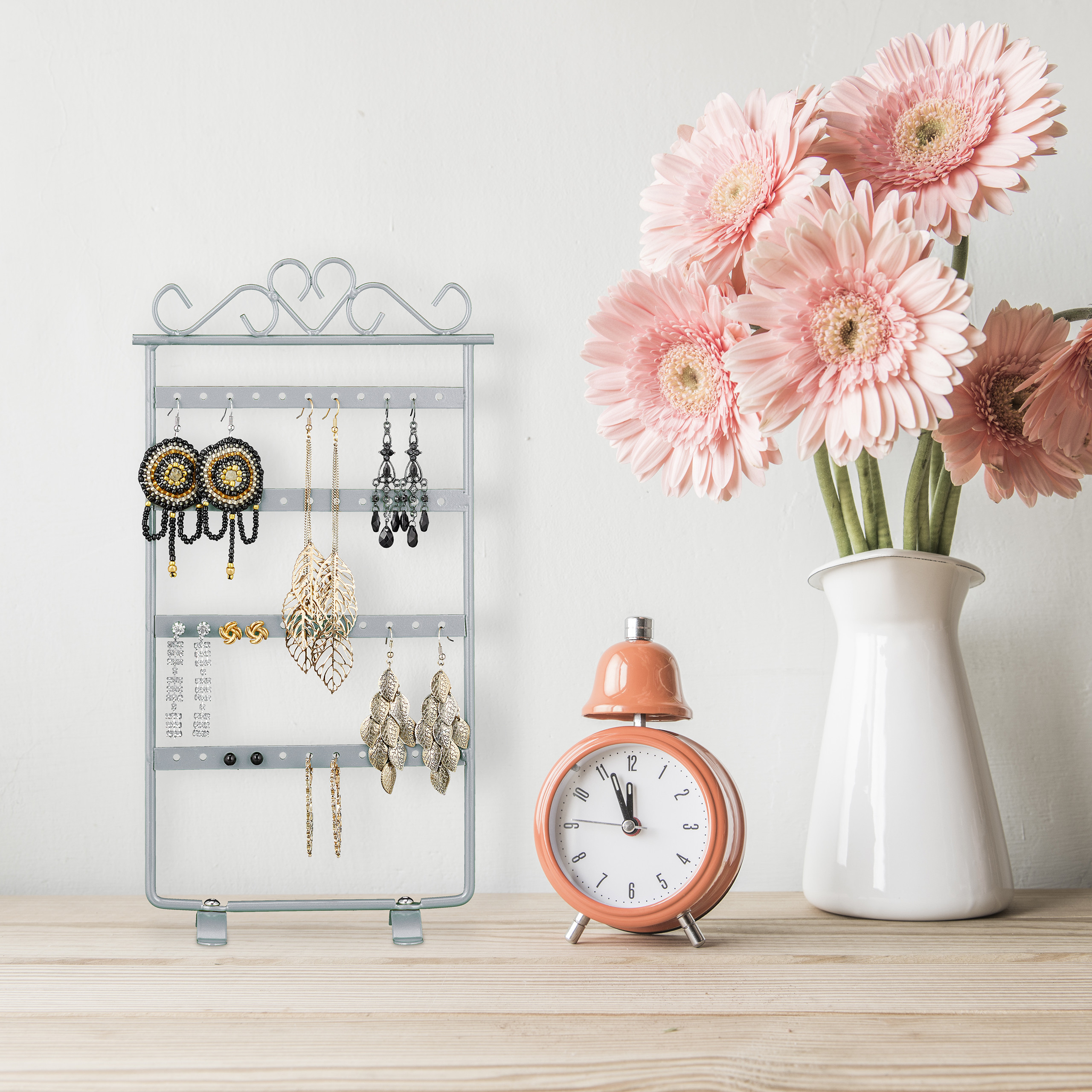 Ein Ohrringständer aus Metall, ein oranger Wecker und eine weiße Vase mit rosa Gerbera auf Holz.