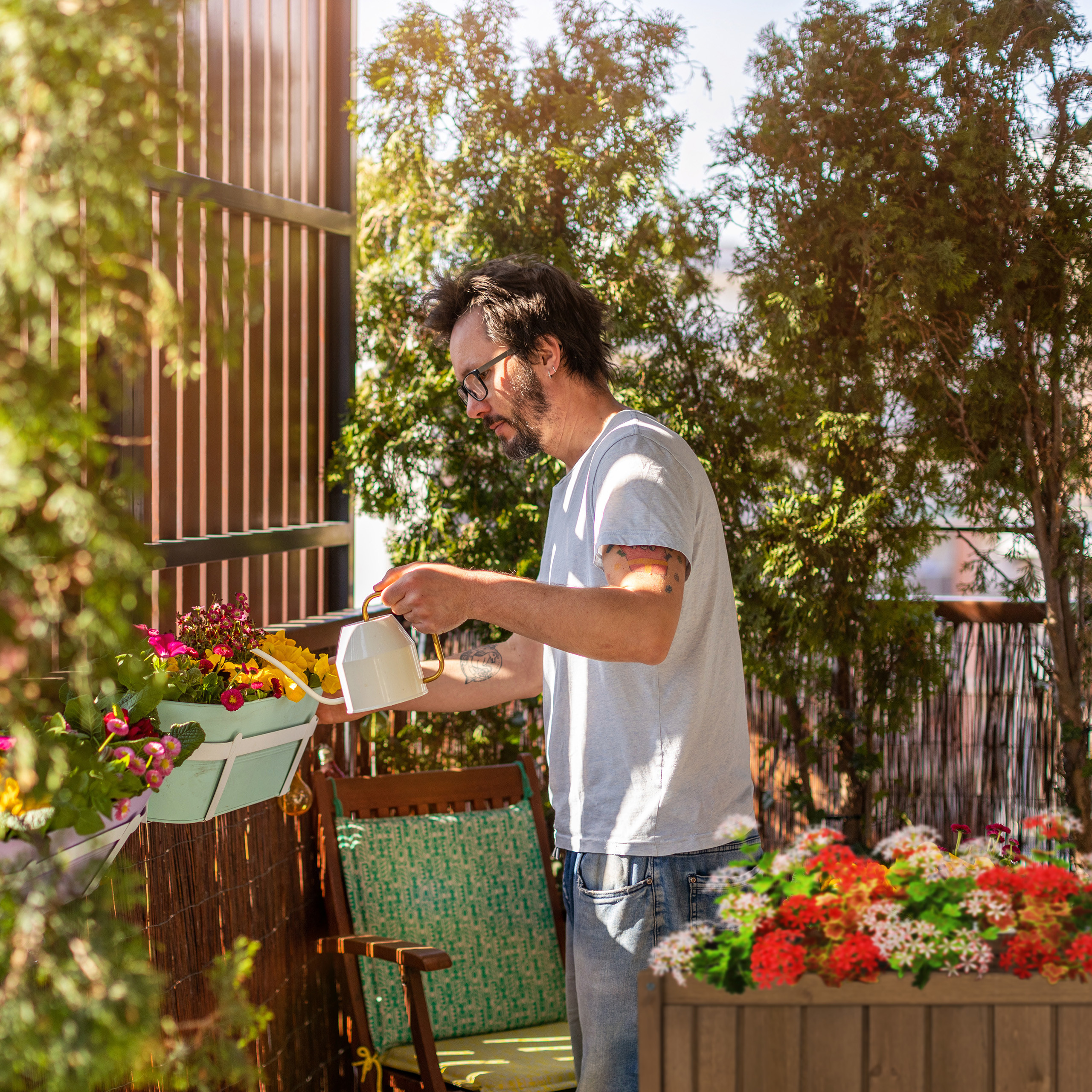 Ein Mann im hellen Hemd gießt Blumen auf einem sonnigen Balkon mit grünen Pflanzen im Hintergrund.