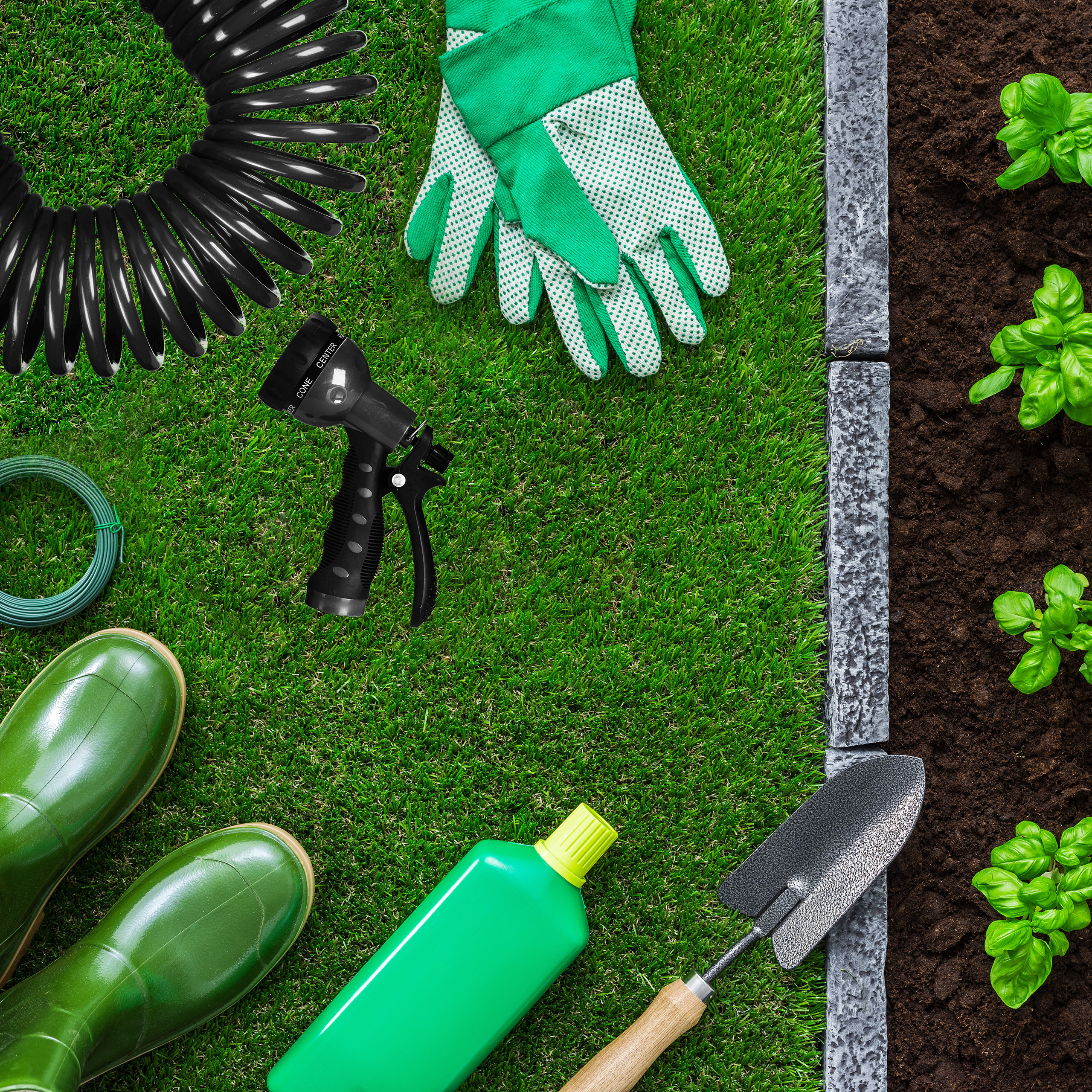 Gartenzubehör wie Stiefel, Handschuhe, Schlauch und Schaufel auf Rasen neben einem Beet.