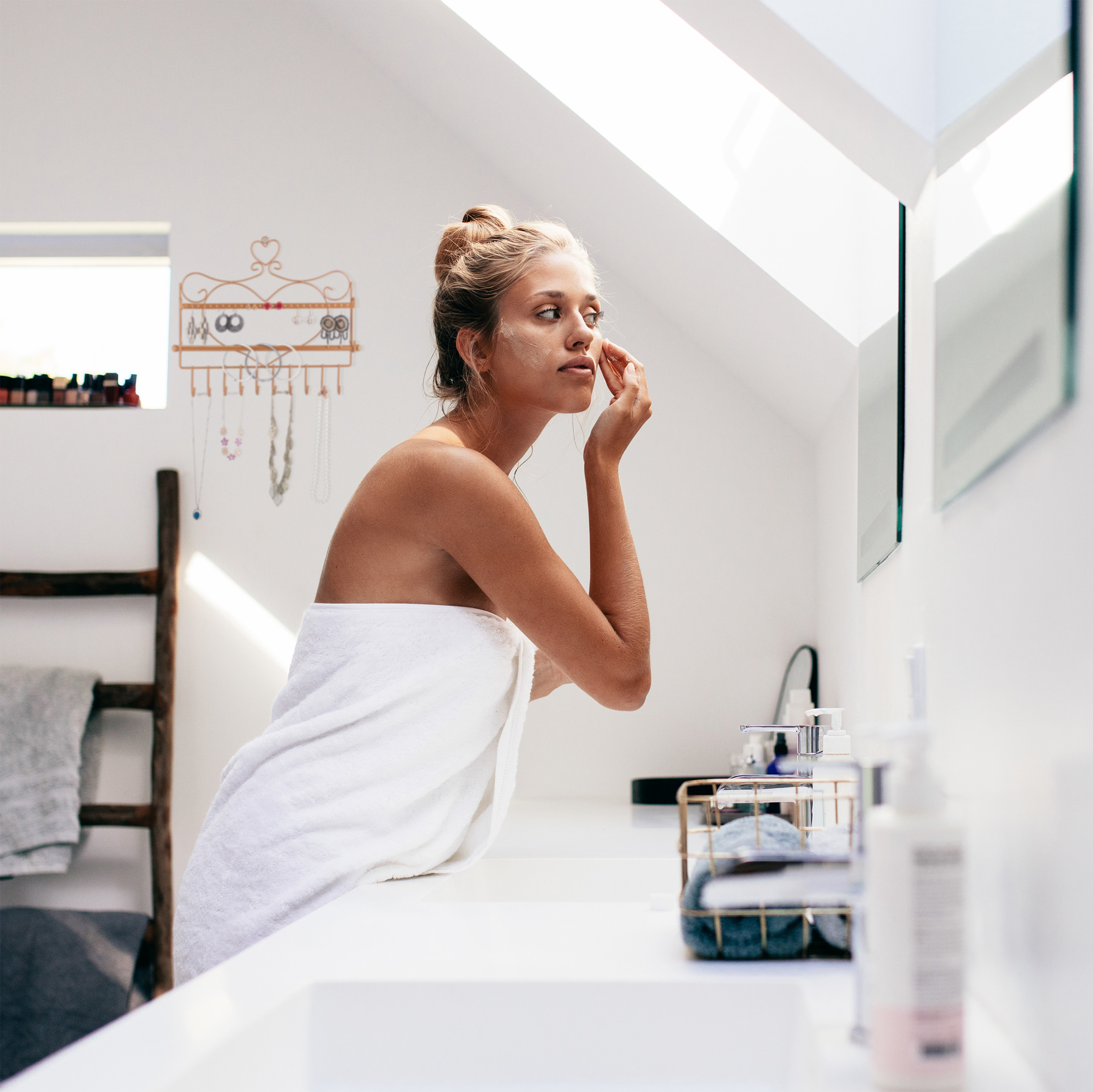 Eine blonde Frau im weißen Handtuch trägt Creme auf ihr Gesicht in einem hellen, modernen Bad auf.