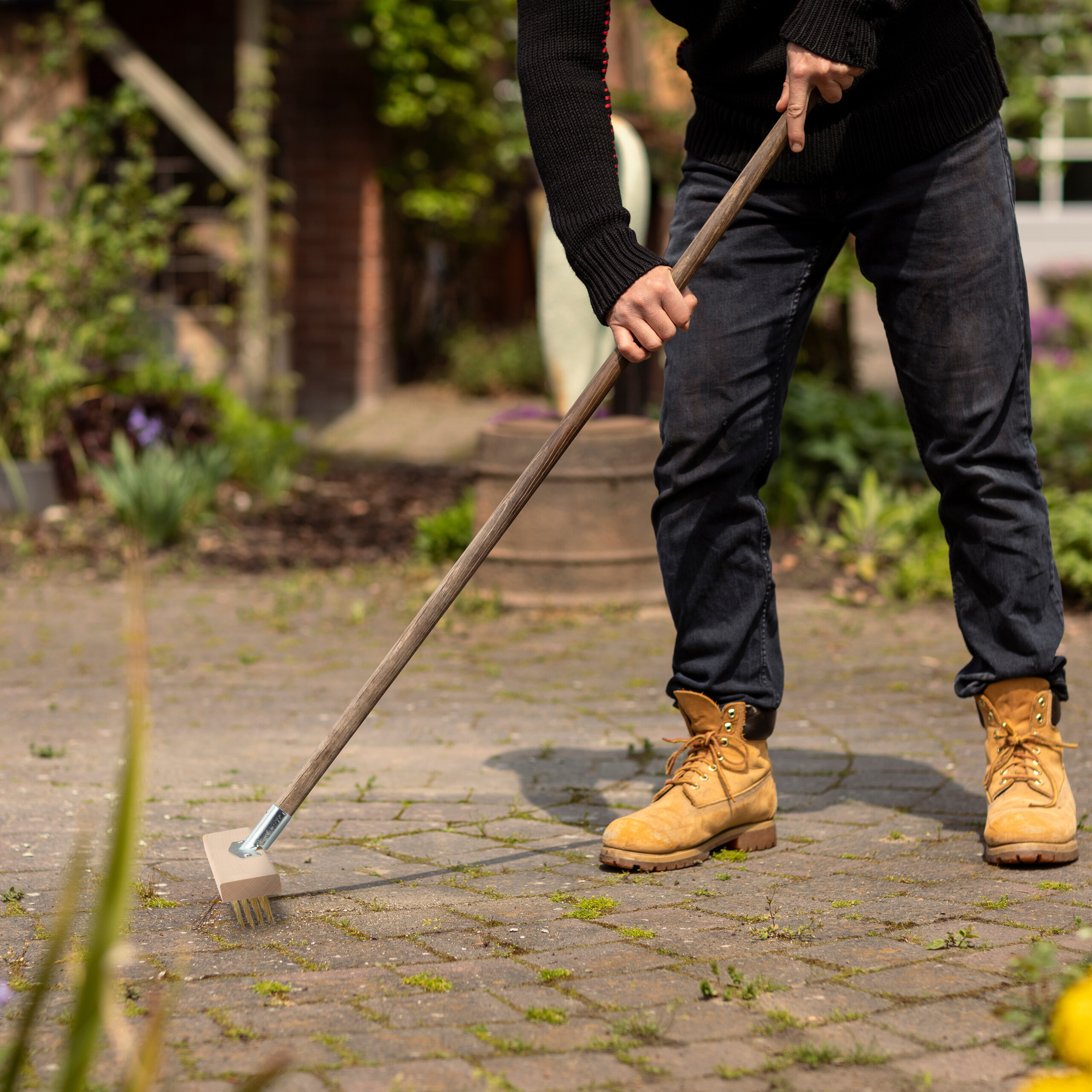 Person in dunkler Kleidung und gelben Stiefeln fegt mit einem Schrubber einen gepflasterten Gartenweg.