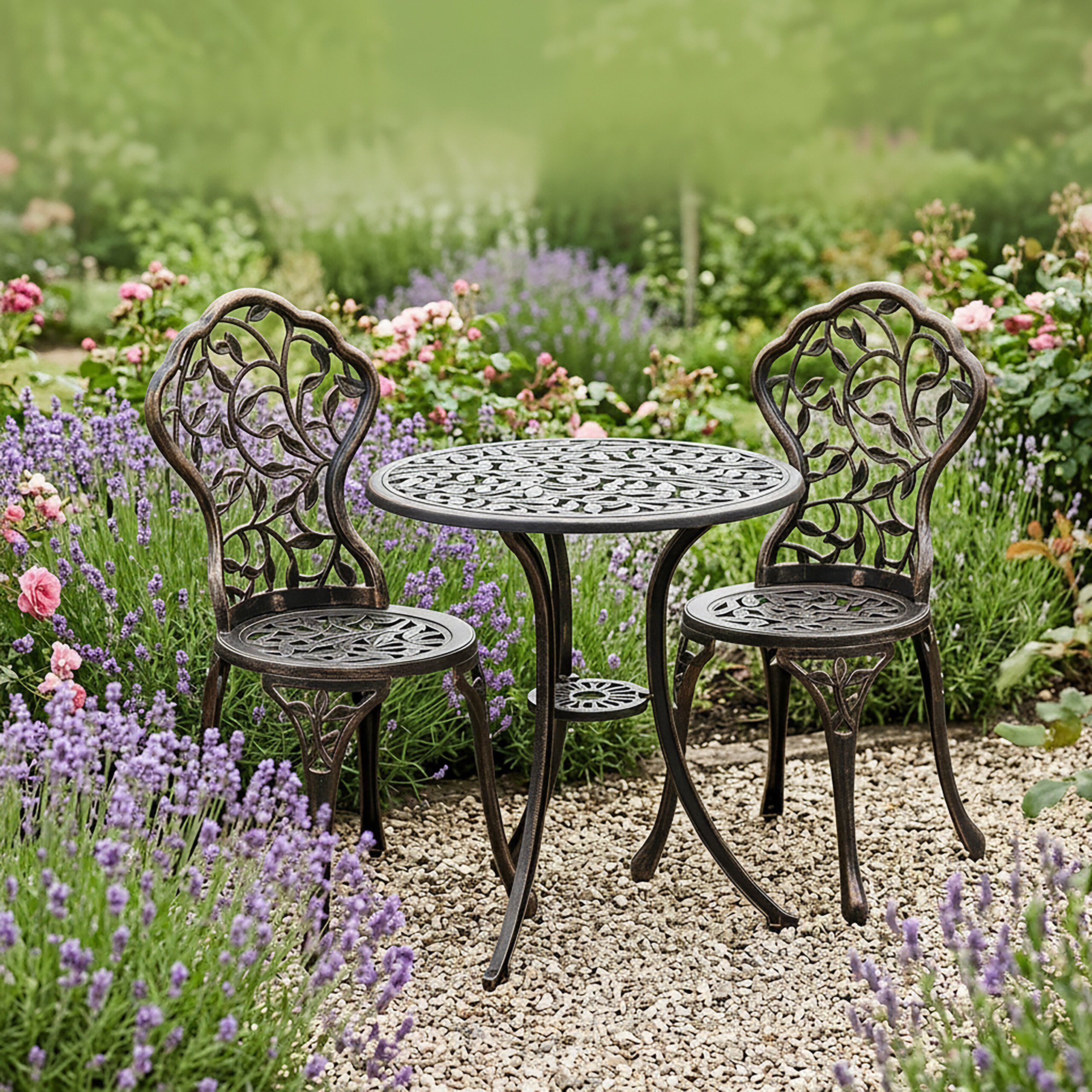 Ein dunkler Metall-Bistrotisch und zwei Stühle stehen auf einem Kiesweg umgeben von blühendem Lavendel und Blumen.