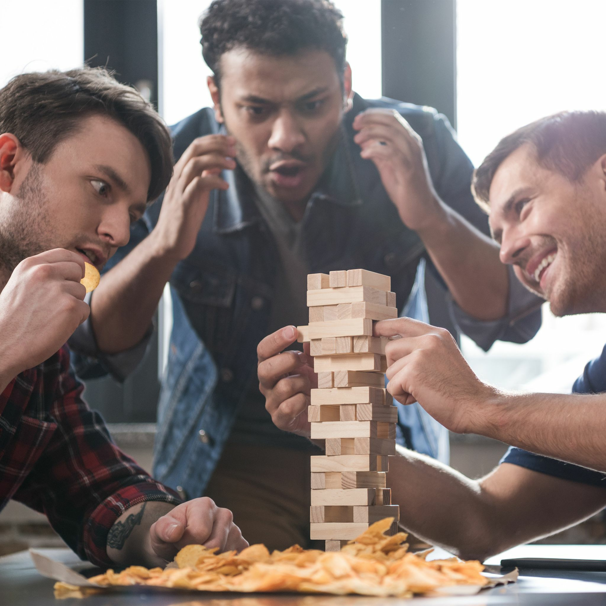 Drei Männer spielen Jenga, ein Mann isst Chips und schaut angespannt zu.