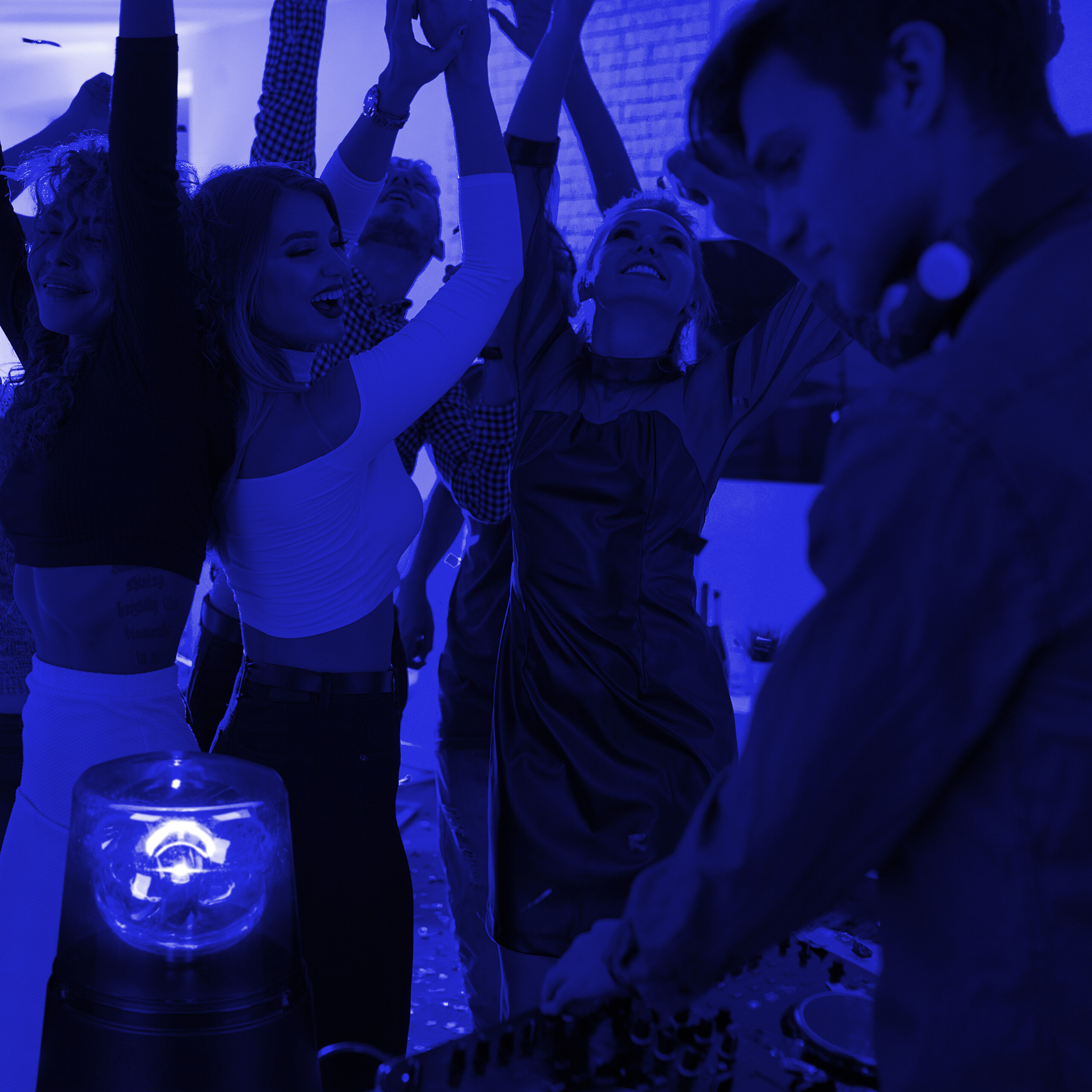 Menschen tanzen mit erhobenen Händen in einem Club mit blauem Licht und einem DJ im Vordergrund.