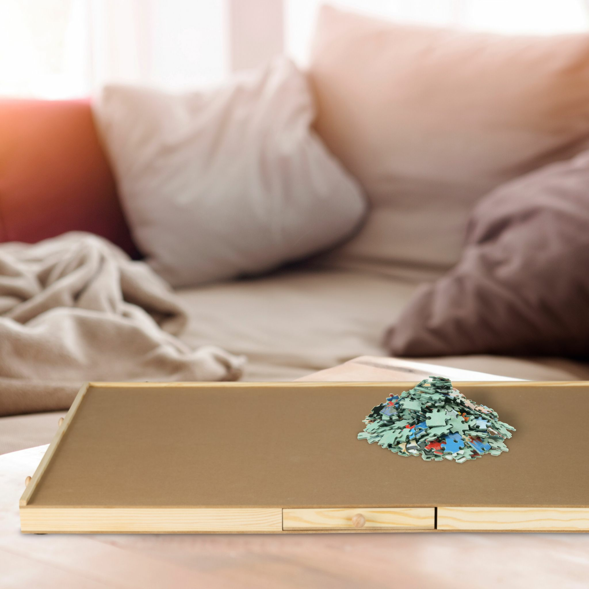 Ein Haufen bunter Puzzleteile liegt auf einem Holzbrett vor einer verschwommenen Couch mit Kissen.