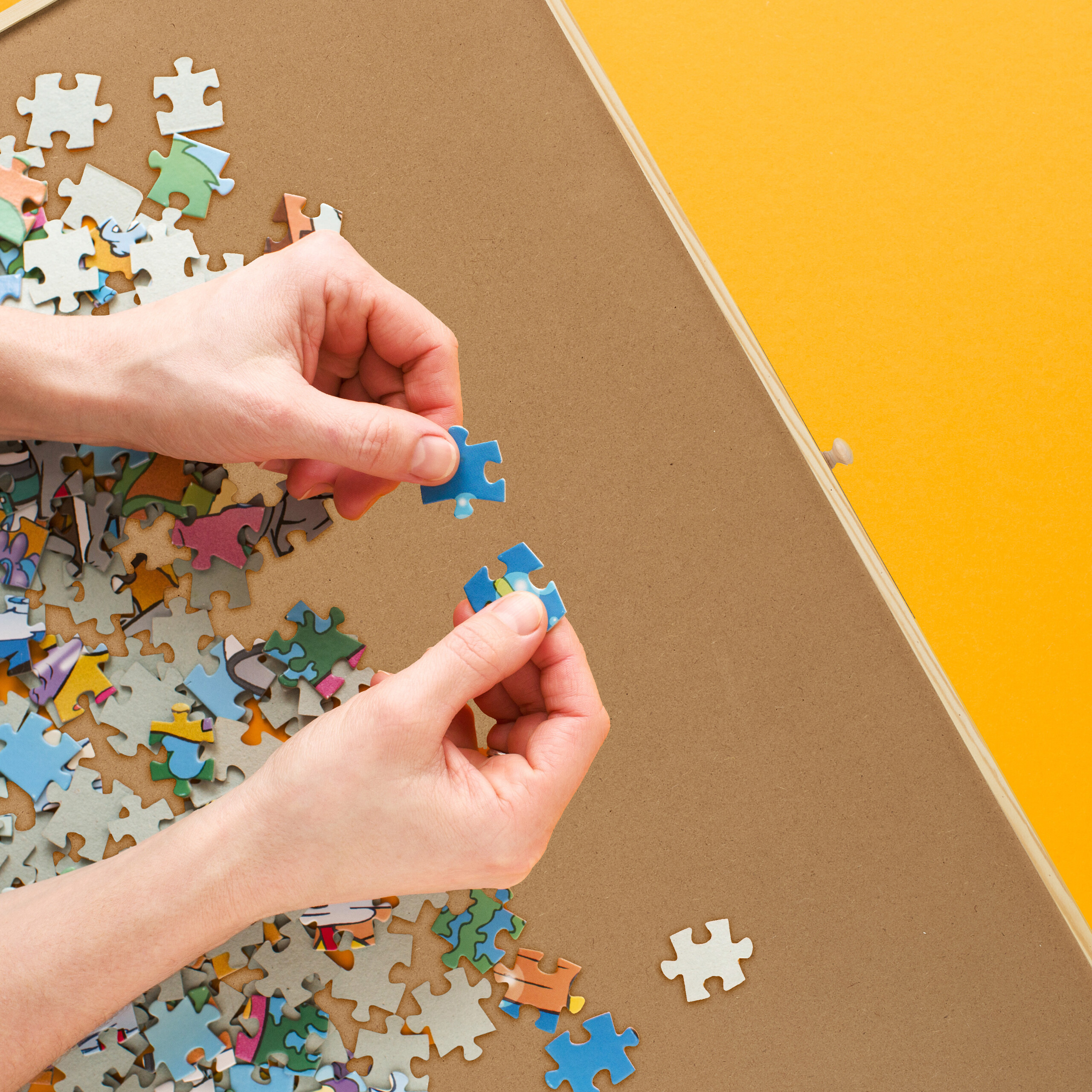 Zwei Hände setzen ein bläuliches Puzzleteil auf einer Holzplatte ein, umgeben von vielen bunten Teilen.