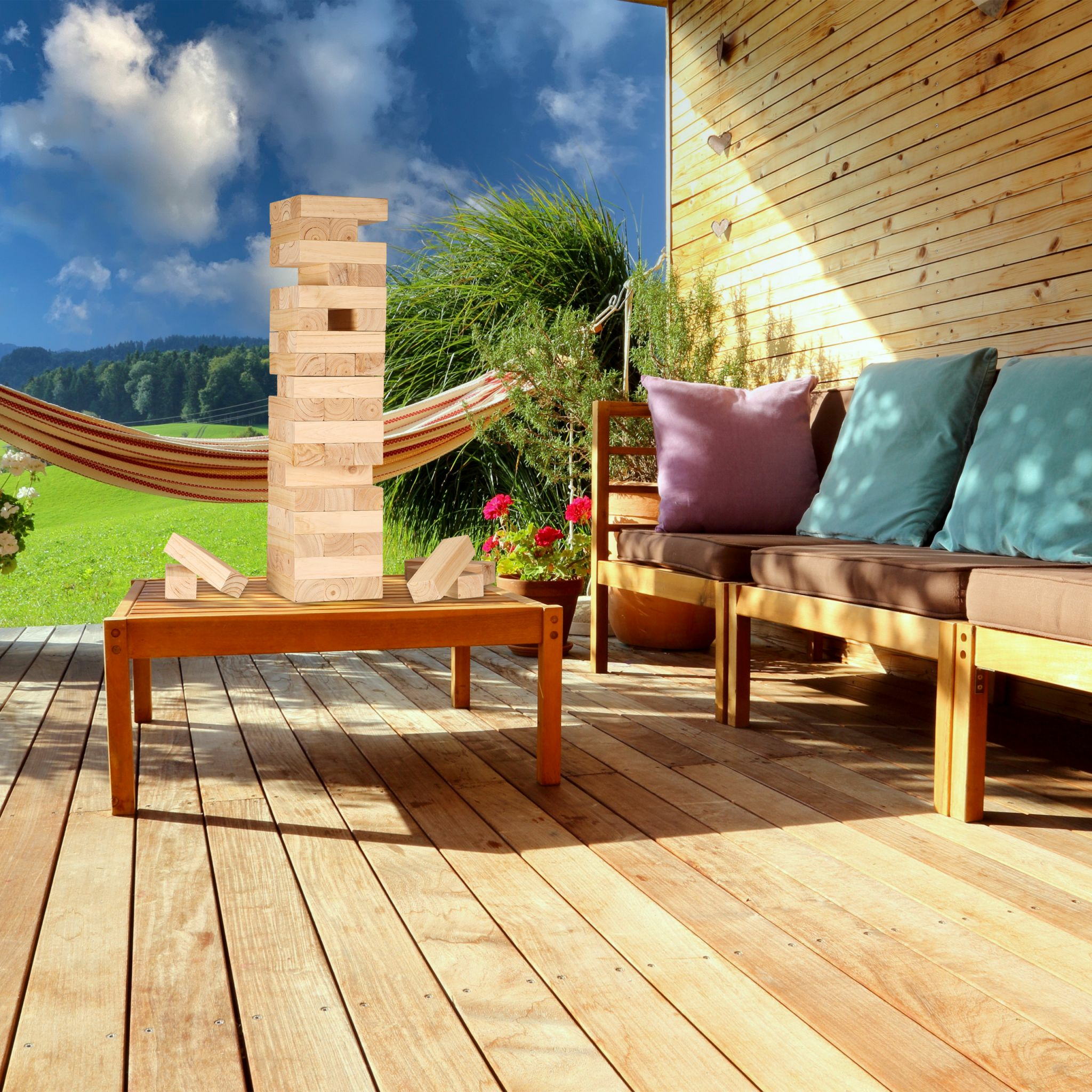 Ein Holzturm steht auf einem Tisch auf einer sonnigen Terrasse mit Sofa, Hängematte und grüner Landschaft.