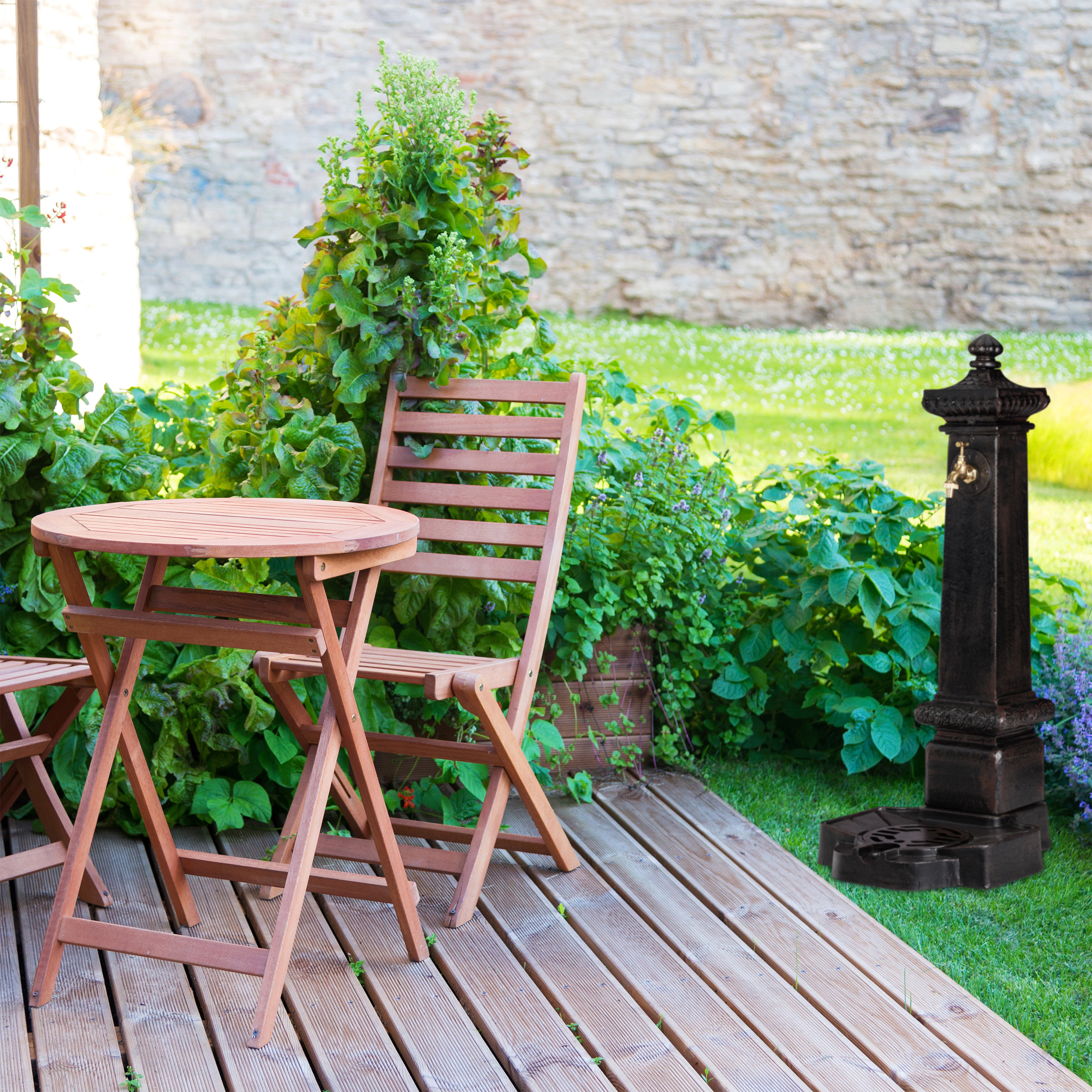 Ein Holztisch und Stühle auf einer Terrasse mit einem dunklen Außenbrunnen, ü Grün und einer Steinmauer im Hintergrund.