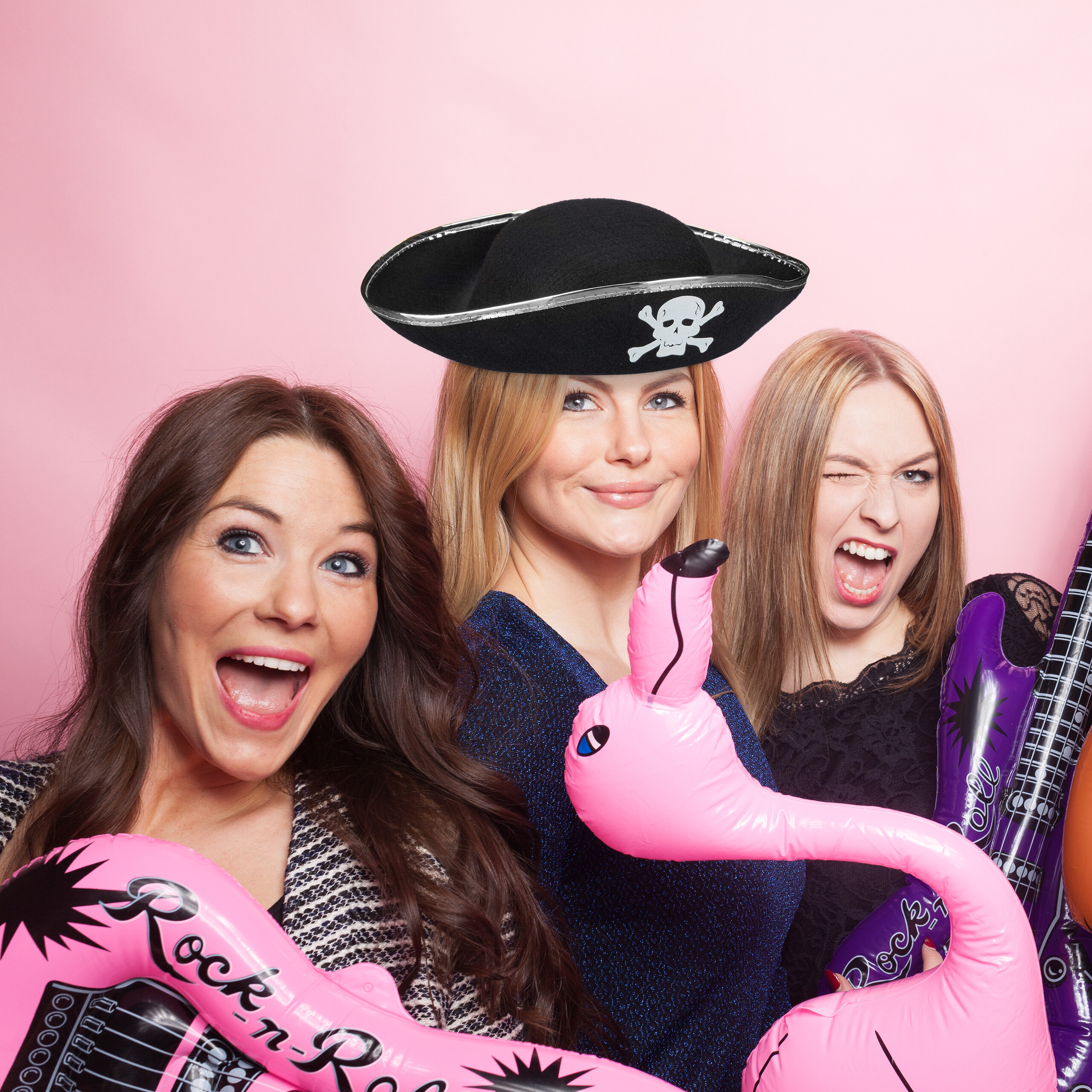 Drei Frauen posieren vor rosa Hintergrund mit Piratenhut und aufblasbaren Gitarren.