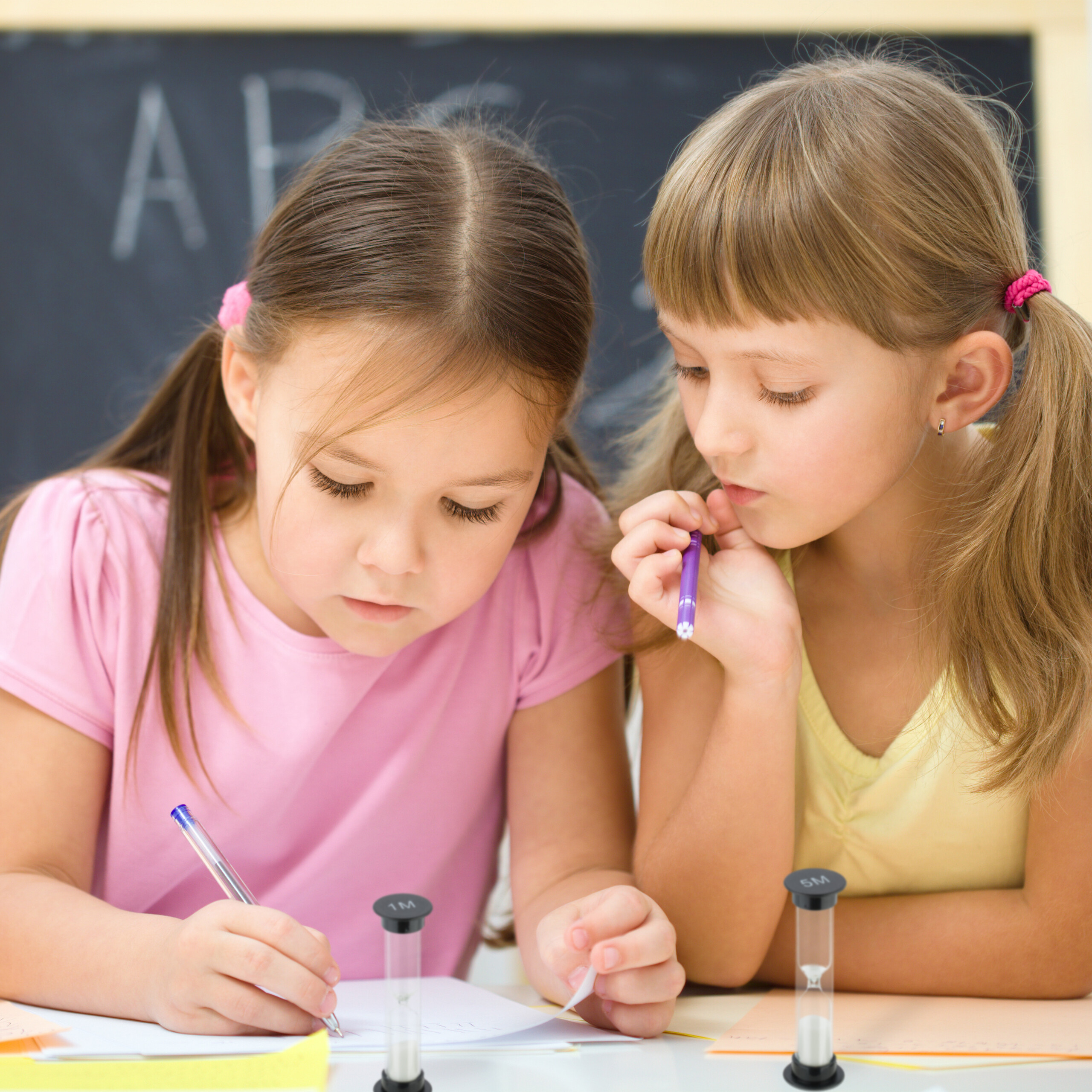 Zwei kleine Mädchen in bunten Hemden schreiben an einem Schreibtisch vor einer Schultafel.