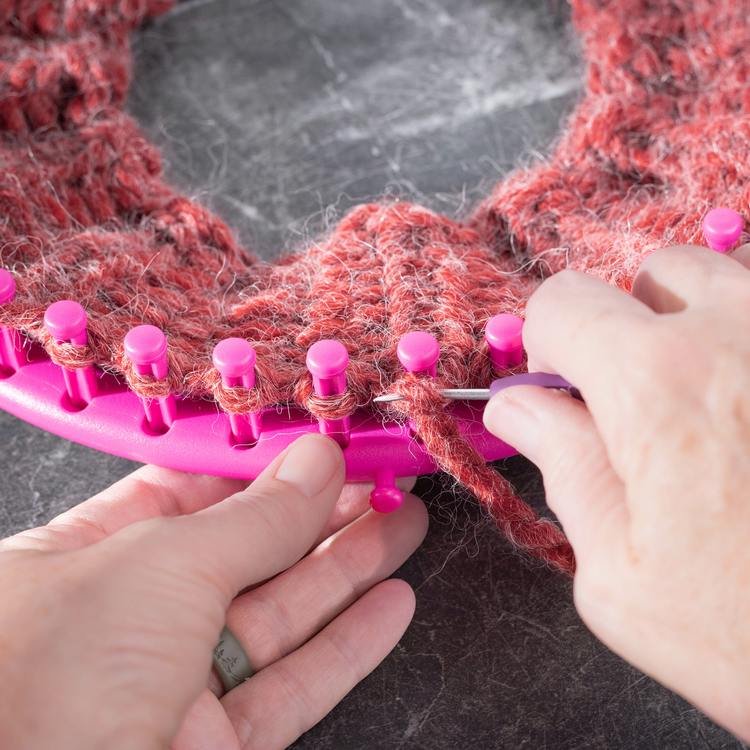 Hände nutzen eine Häkelnadel an einem rosa Strickrahmen mit roter Wolle.