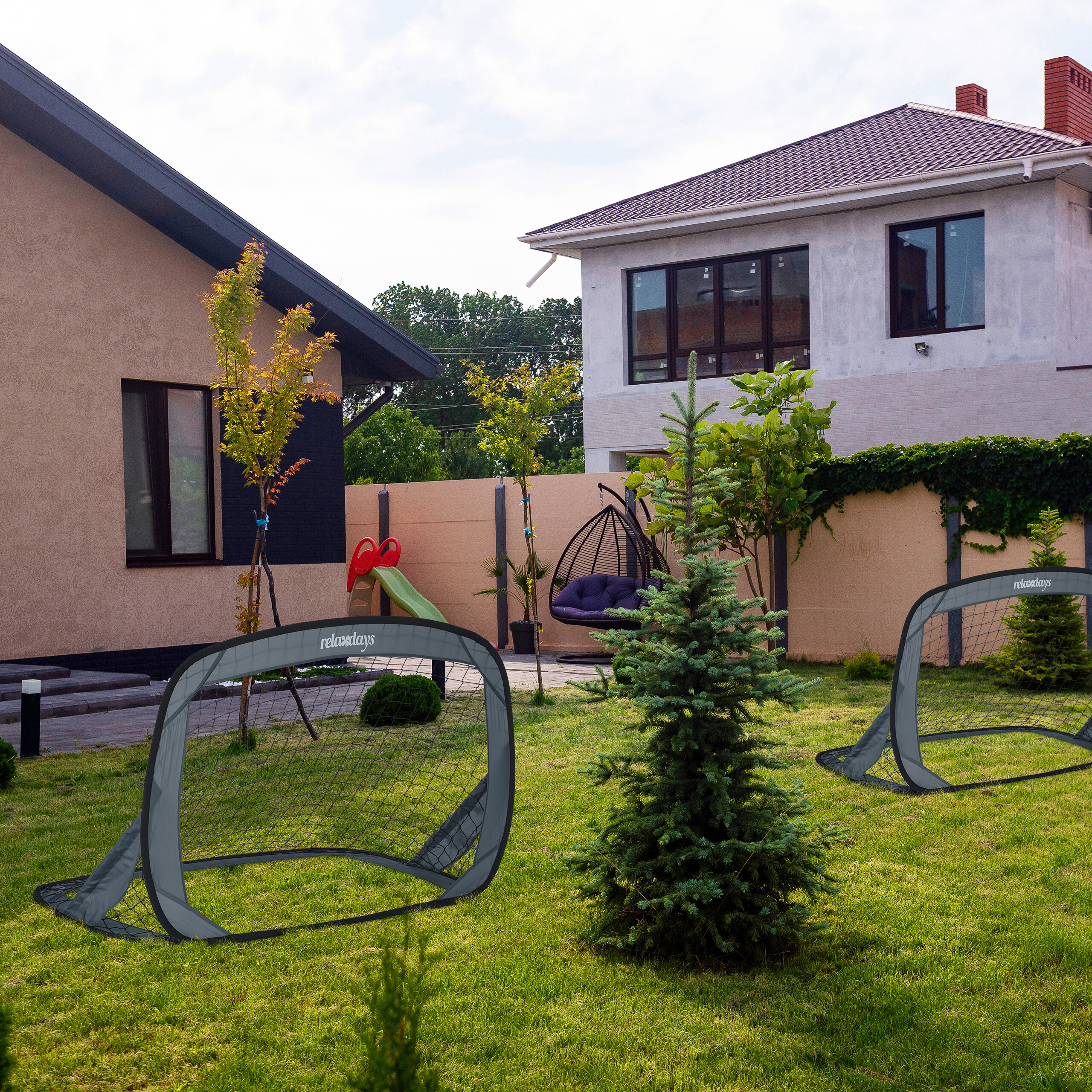 Zwei Pop-up-Fußballtore auf einem grünen Rasen in einem Vorstadthof mit Häusern, Bäumen und Hängesessel.
