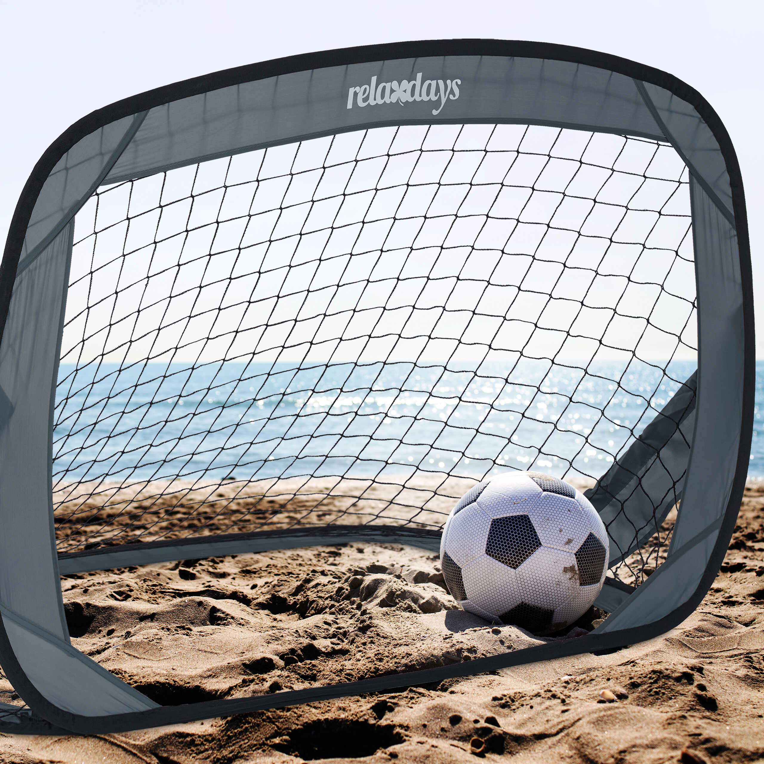 Ein Pop-up-Fußballtor steht am Sandstrand vor dem blauen Meer, ein Fußball liegt im Tor.