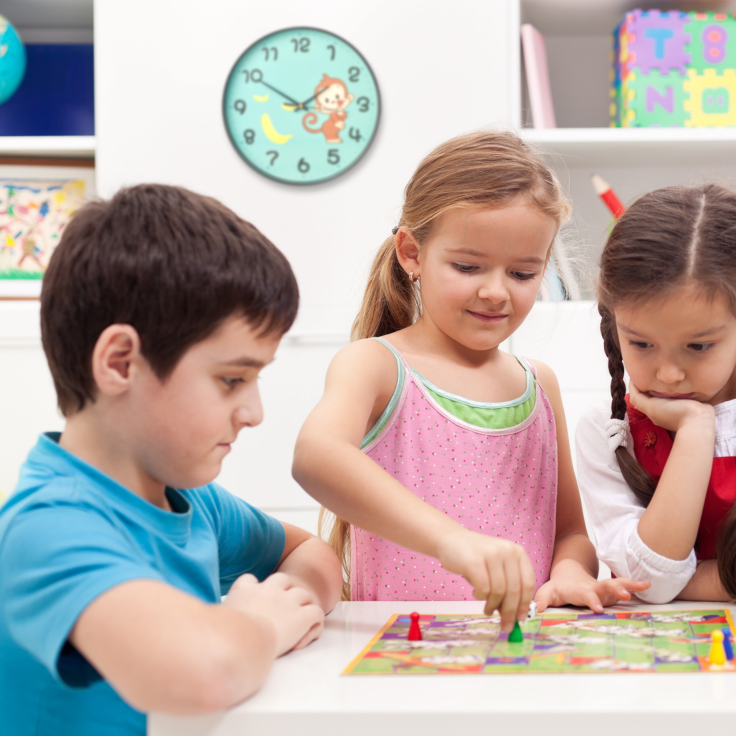 Drei Kinder sitzen an einem Tisch und spielen ein farbiges Brettspiel, an der Wand hängt eine Affenuhr.