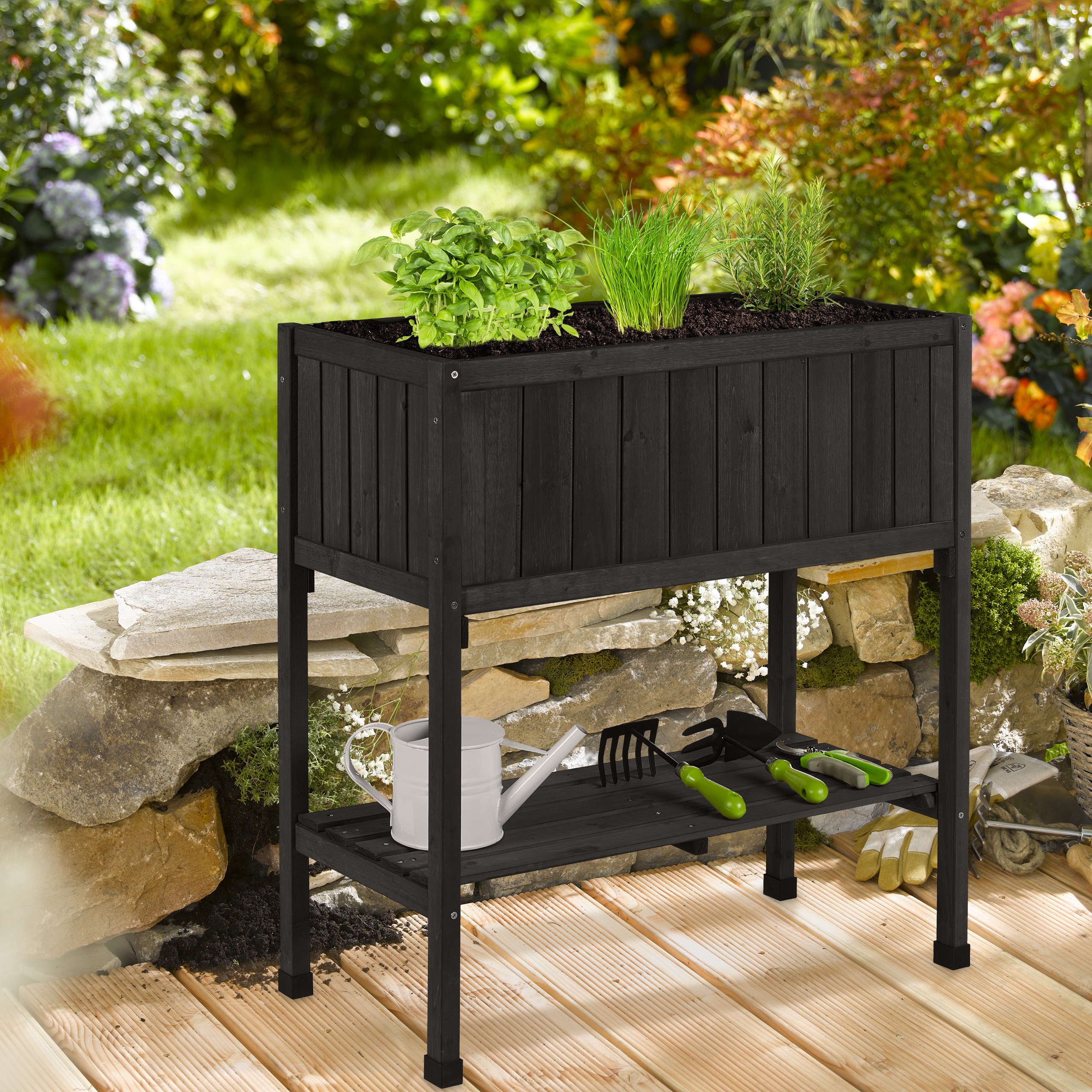 Ein schwarzes Hochbeet aus Holz mit Kräutern, darunter Gießkanne und Werkzeug, auf einer Holzterrasse.