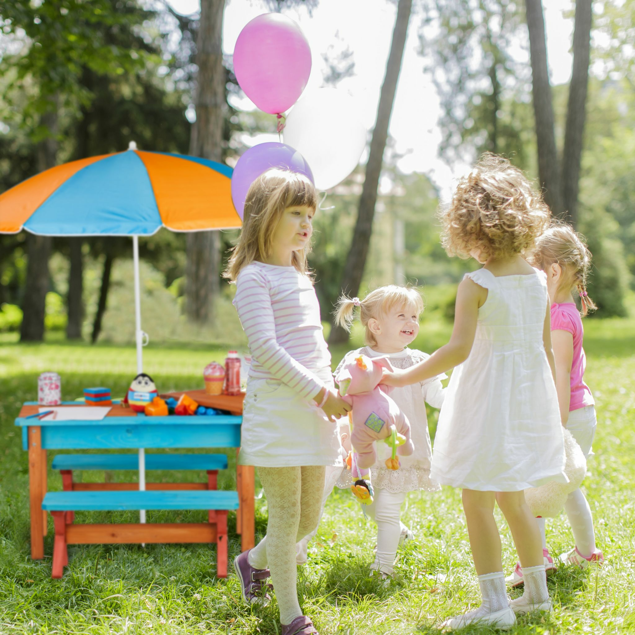 Vier junge Mädchen spielen in einem sonnigen Park mit bunten Luftballons, einem Picknicktisch und einem Schirm.